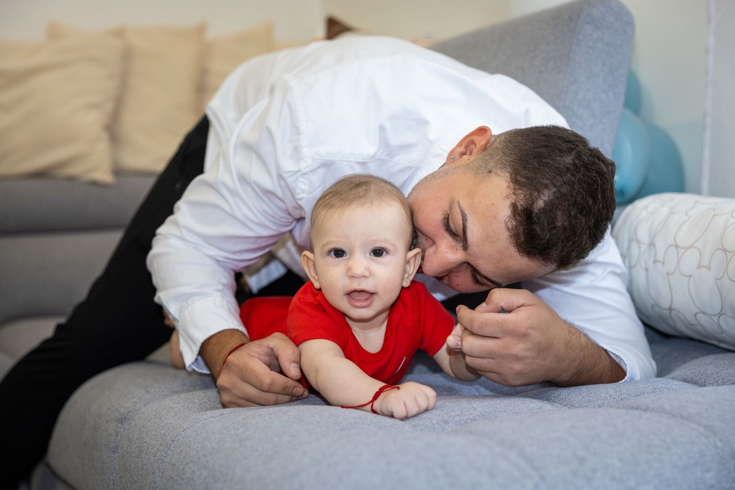 A loving father in a white shirt kisses his baby in a red onesie while lying on a gray sofa.