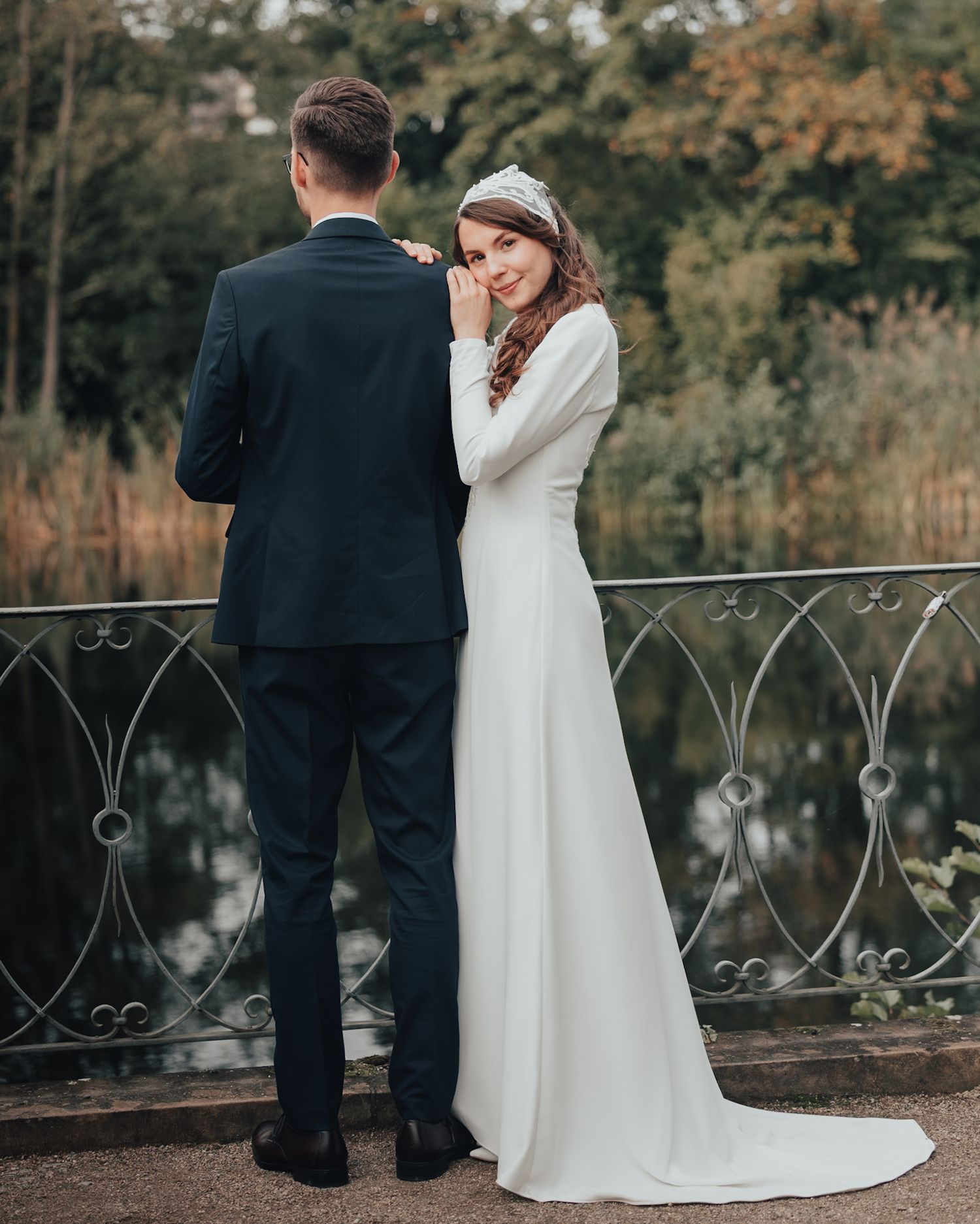 Bride and groom standing together by ornate railing overlooking water with autumn foliage in background.