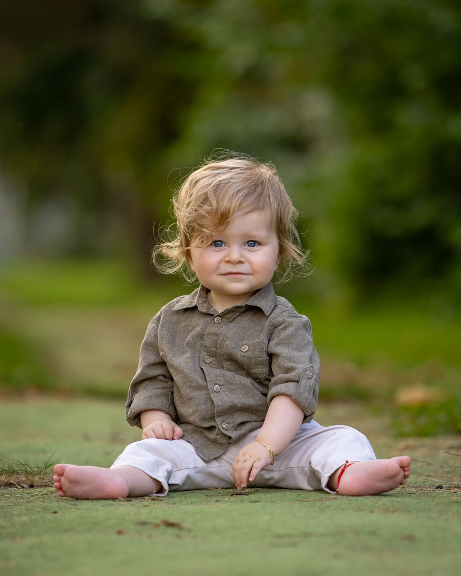 Adorable toddler with curly blonde hair sits cross-legged on grass, wearing a olive shirt and white pants.