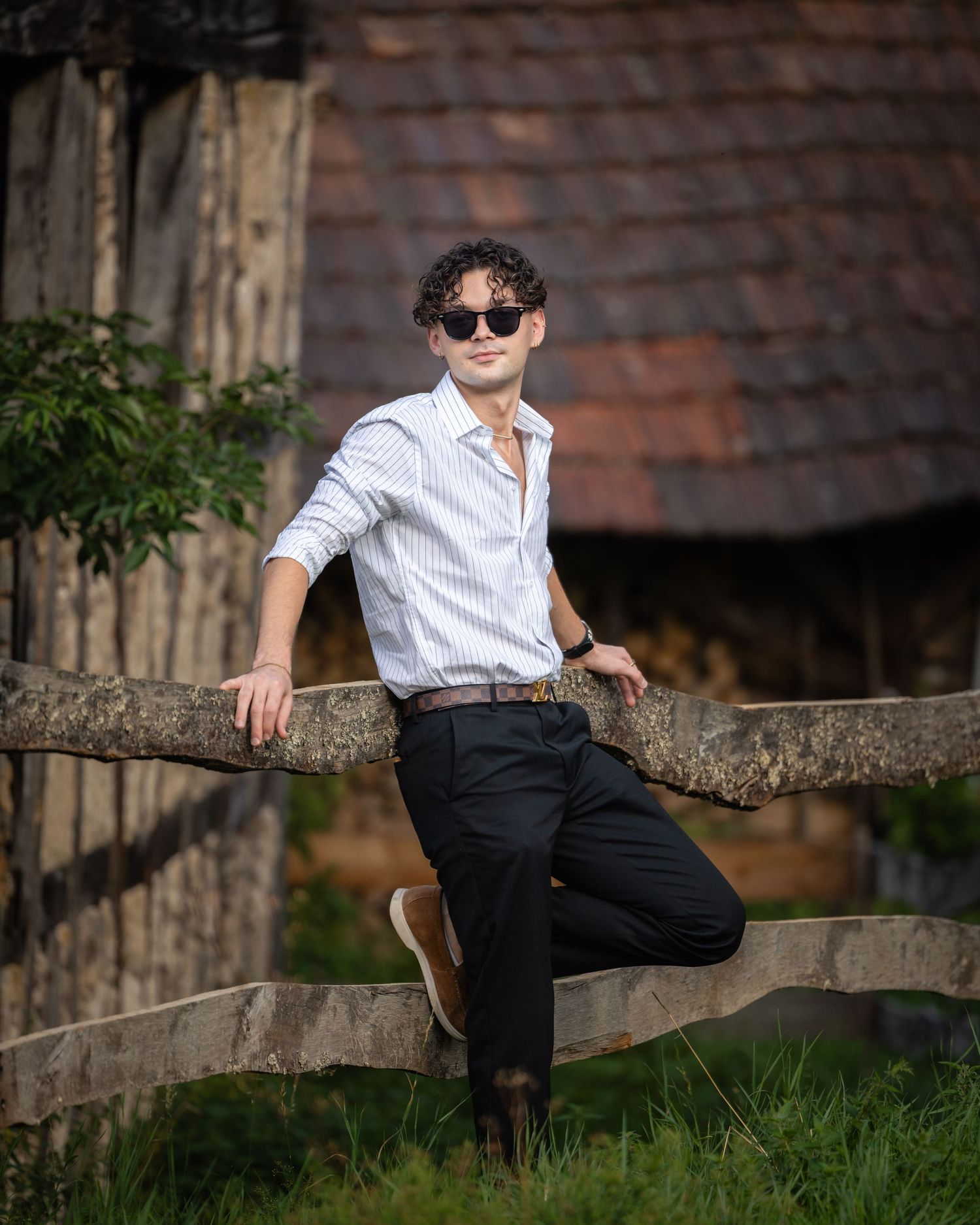 Young man in striped shirt and sunglasses leaning on rustic wooden fence near vintage building.