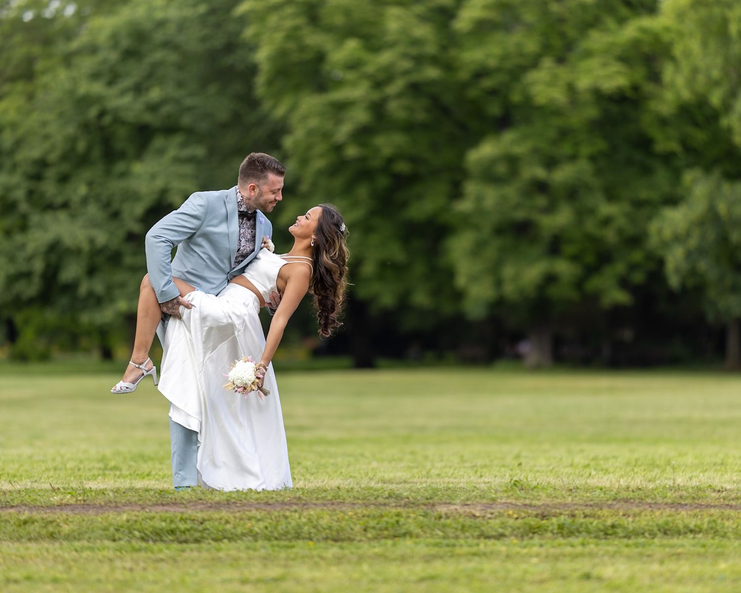 A couple in white clothing embraces joyfully in an open green field with trees in the background.