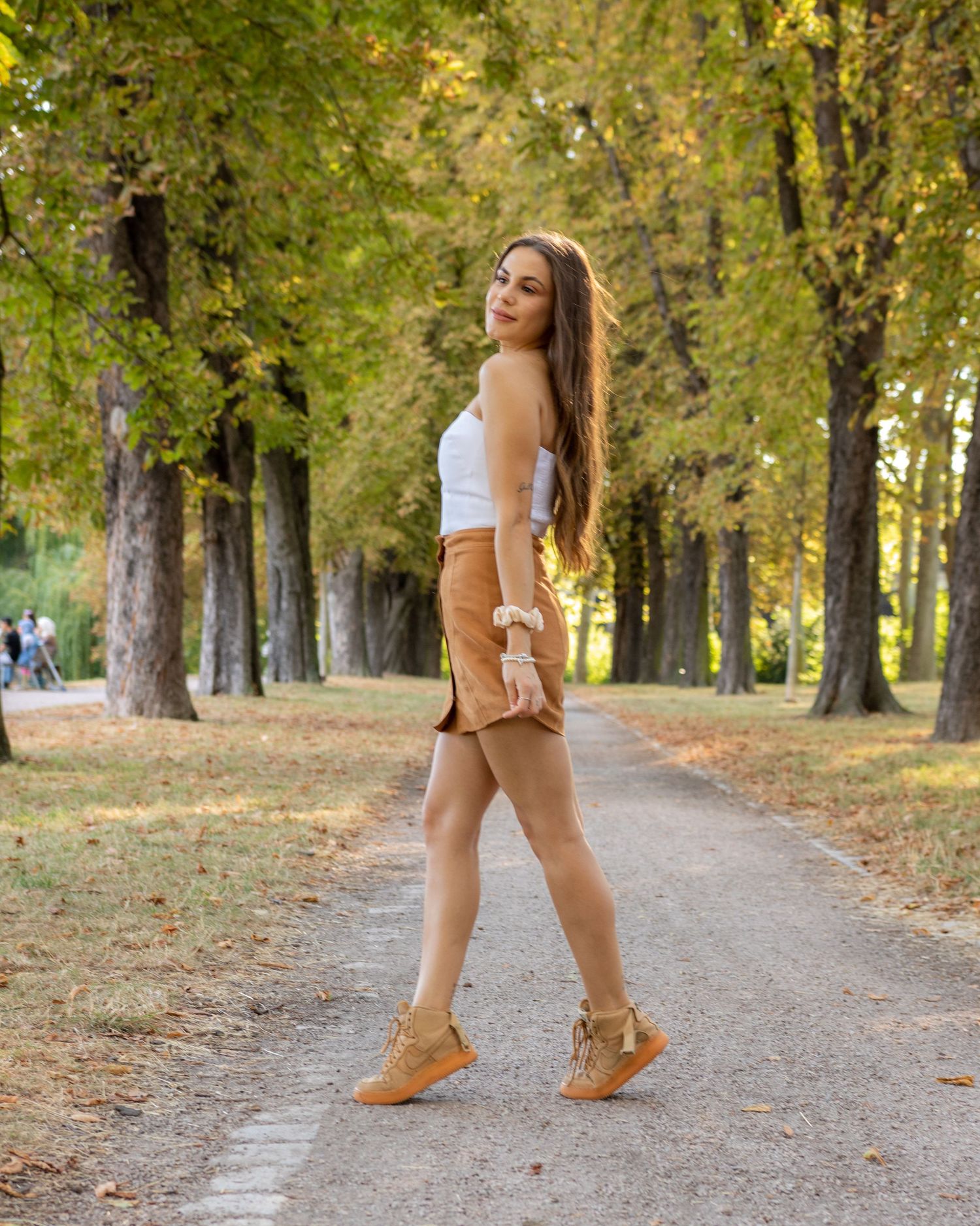Young woman in white top, tan shorts, and fringe boots poses on a tree-lined park path in autumn.