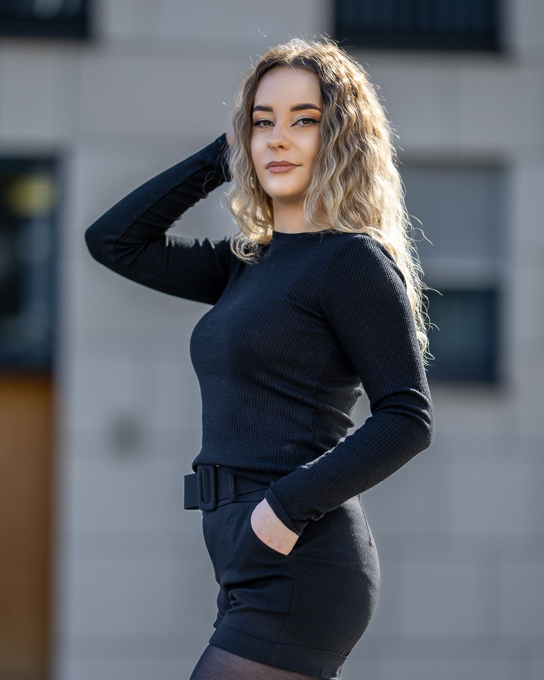 Young woman in black long-sleeve mini dress with belt, posing outdoors with hand in hair.