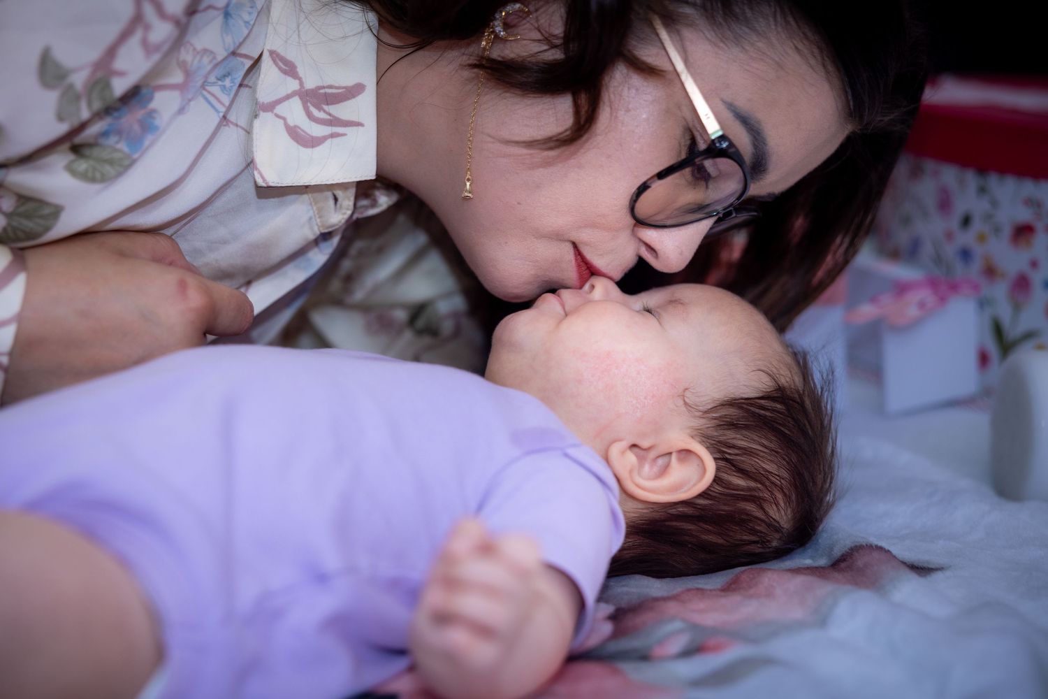 Eine Frau küsst Emilia, die in einem lilafarbenen Outfit auf einer Decke liegt.