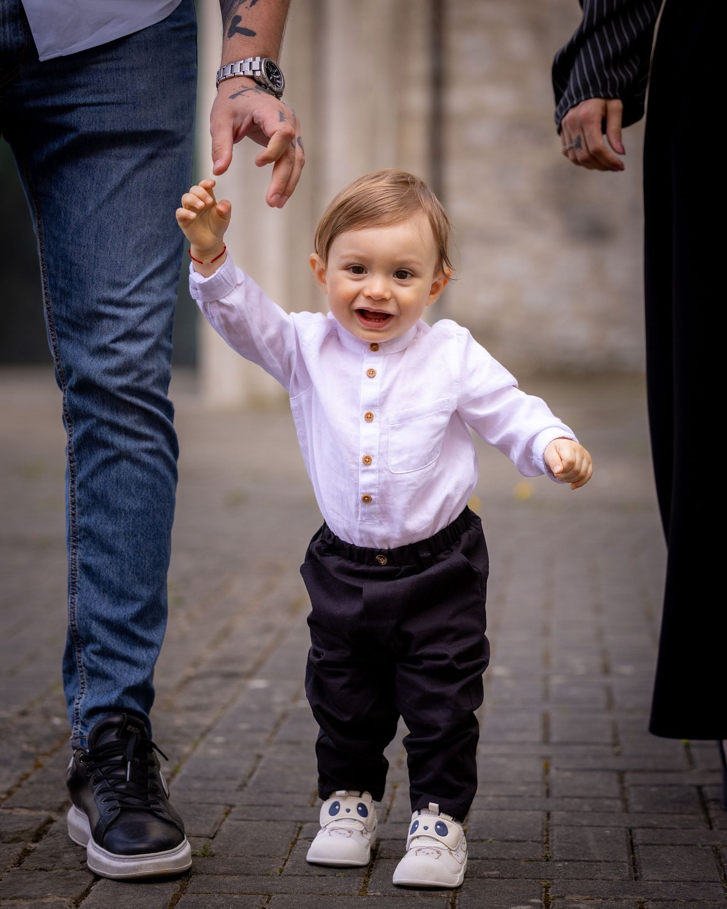 A happy toddler in a white shirt and dark pants holds an adult's hand while learning to walk on a cobblestone path.