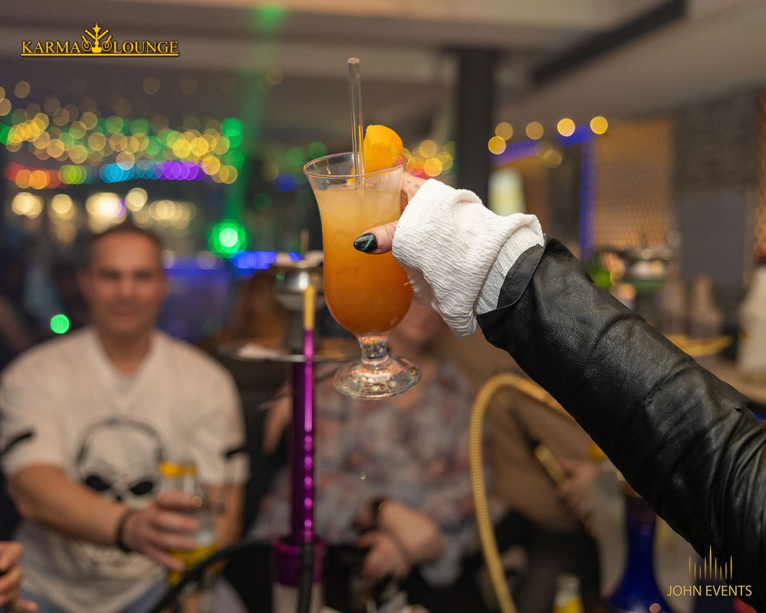 Eine behandschuhte Hand hebt einen orangefarbenen Drink vor den Gästen der Karma Lounge.