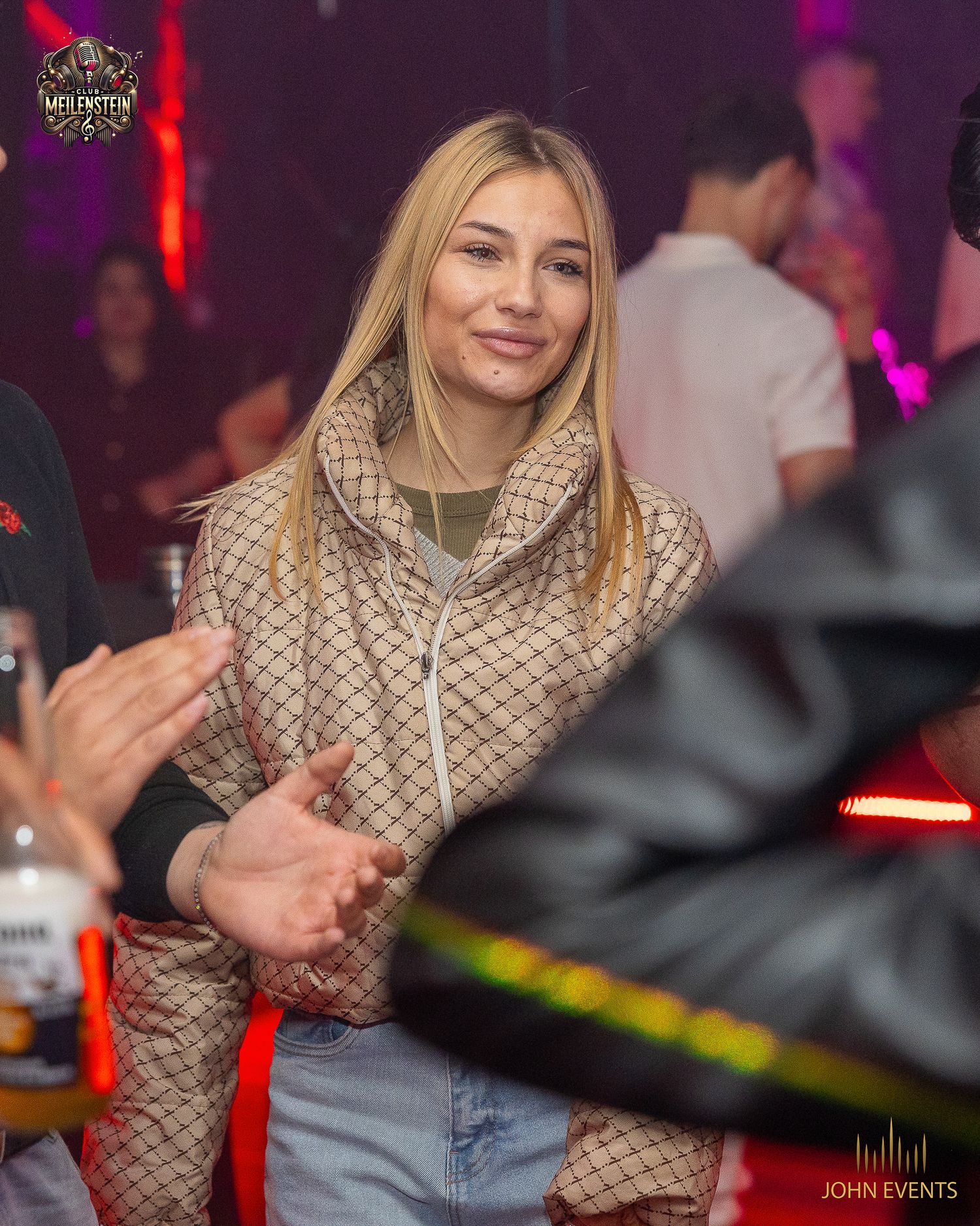Eine blonde Frau mit gemustertem Schal blickt im rot beleuchteten Club Meilenstein zur Kamera.