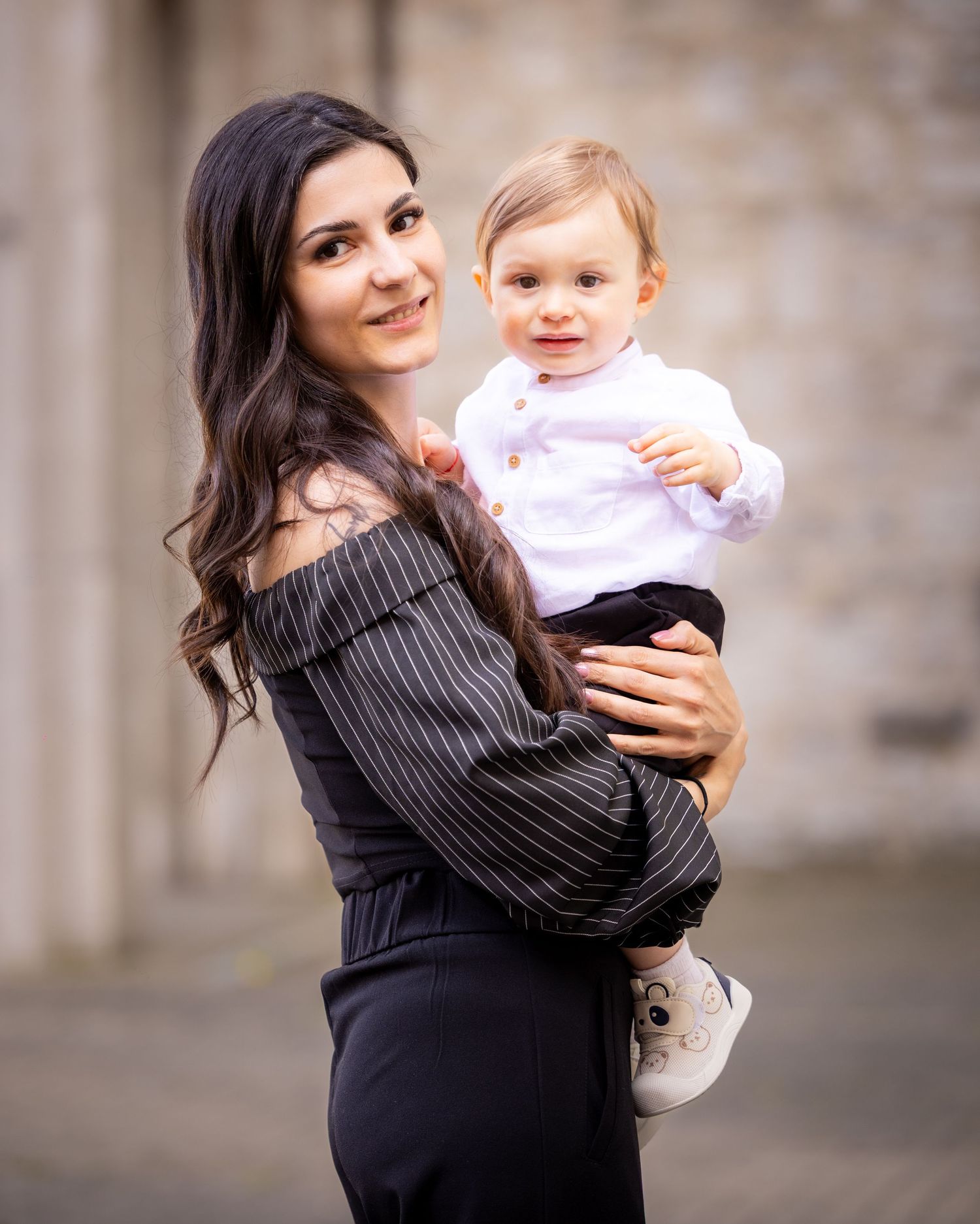 A smiling young woman with long dark hair holds a happy toddler in a white shirt outdoors.