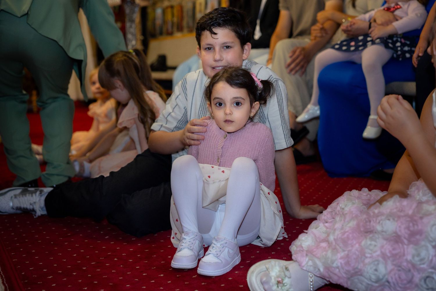 Zwei Kinder sitzen zusammen auf dem roten Teppich, während der ältere Junge das jüngere Mädchen umarmt