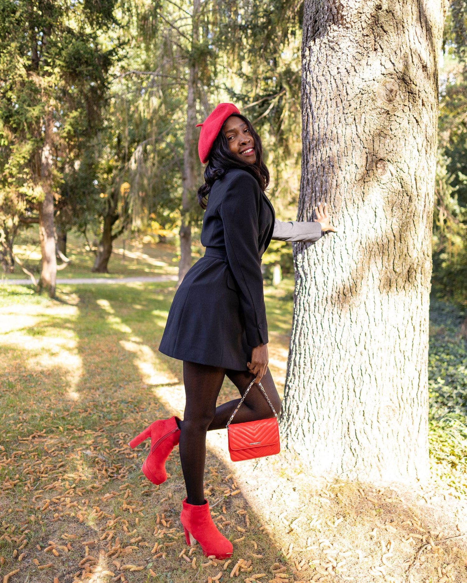 Jacky posiert in schwarzem Kleid mit roten Accessoires an einem großen Baum