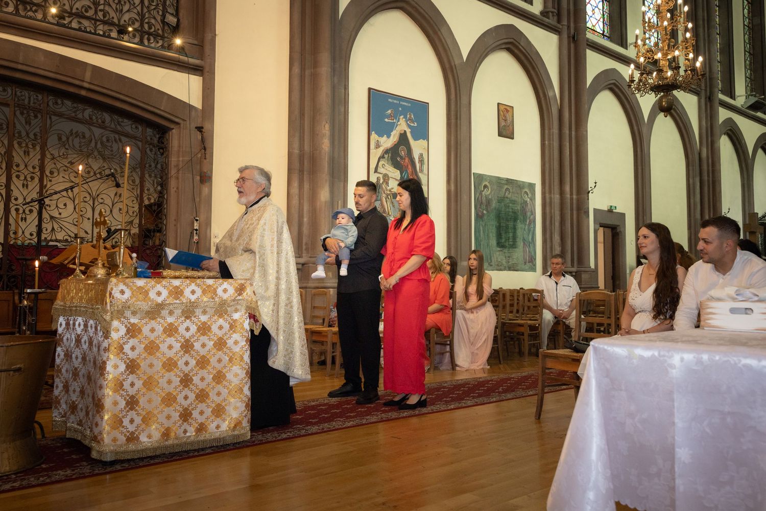 A priest performs a baptism ceremony in an ornate church with family members standing at the altar.