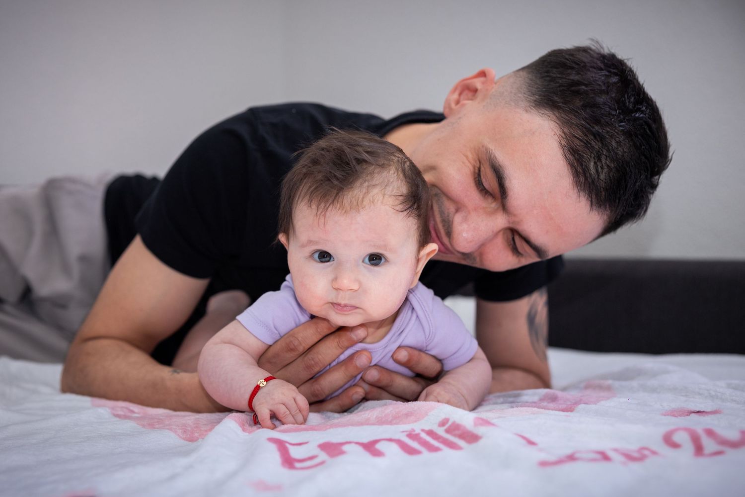 A loving father gently kisses his baby girl lying on a bed with a personalized name blanket.