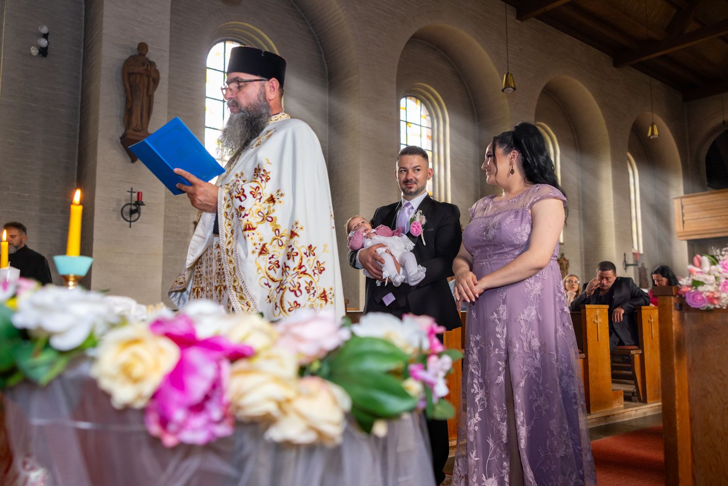 Orthodox priest reads from Bible during infant baptism ceremony with parents holding baby in church.