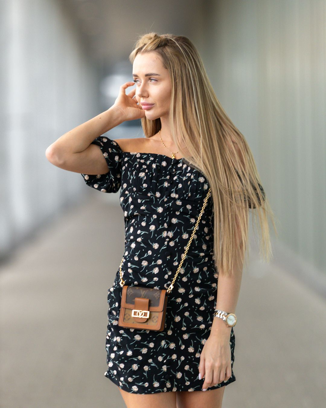 Blonde woman in black floral off-shoulder romper with brown crossbody bag poses in modern corridor.