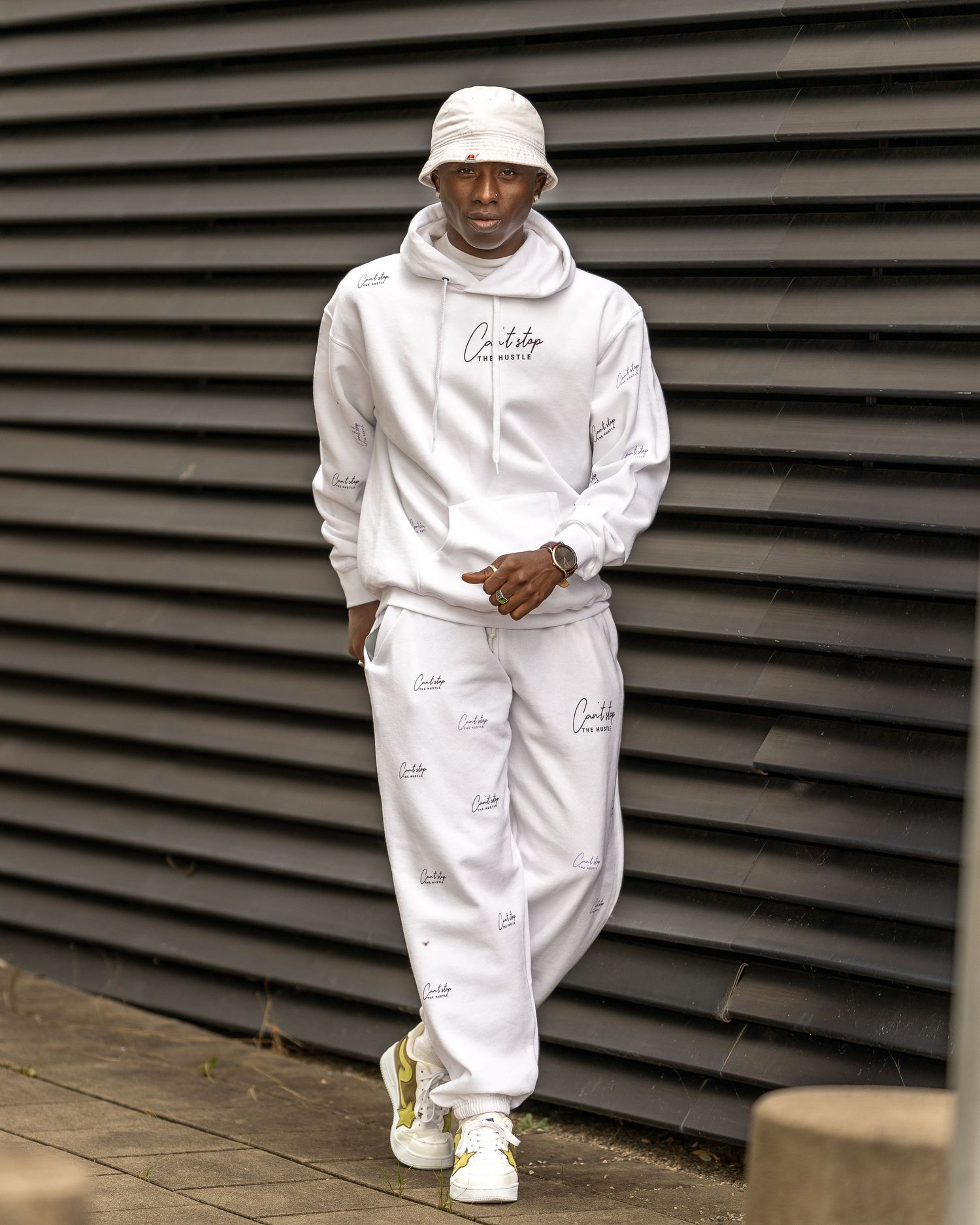 Man wearing white bucket hat, hoodie, and joggers with gold-accented sneakers, posing against dark slatted wall.
