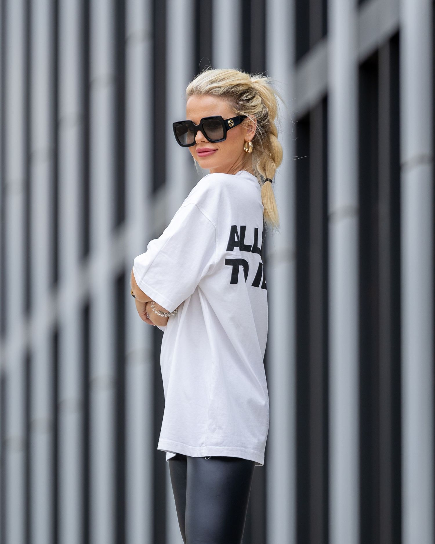 Blonde woman in oversized white graphic tee, black leggings, and sunglasses posing outdoors against striped wall.