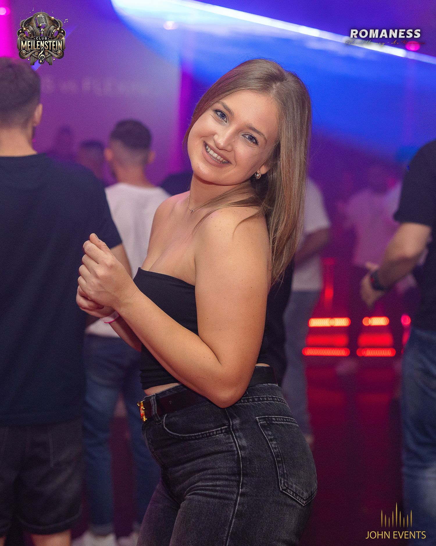 Eine Frau im schwarzen trägerlosen Oberteil und dunkler Jeans lächelt im blau und rot beleuchteten Club Meilenstein.