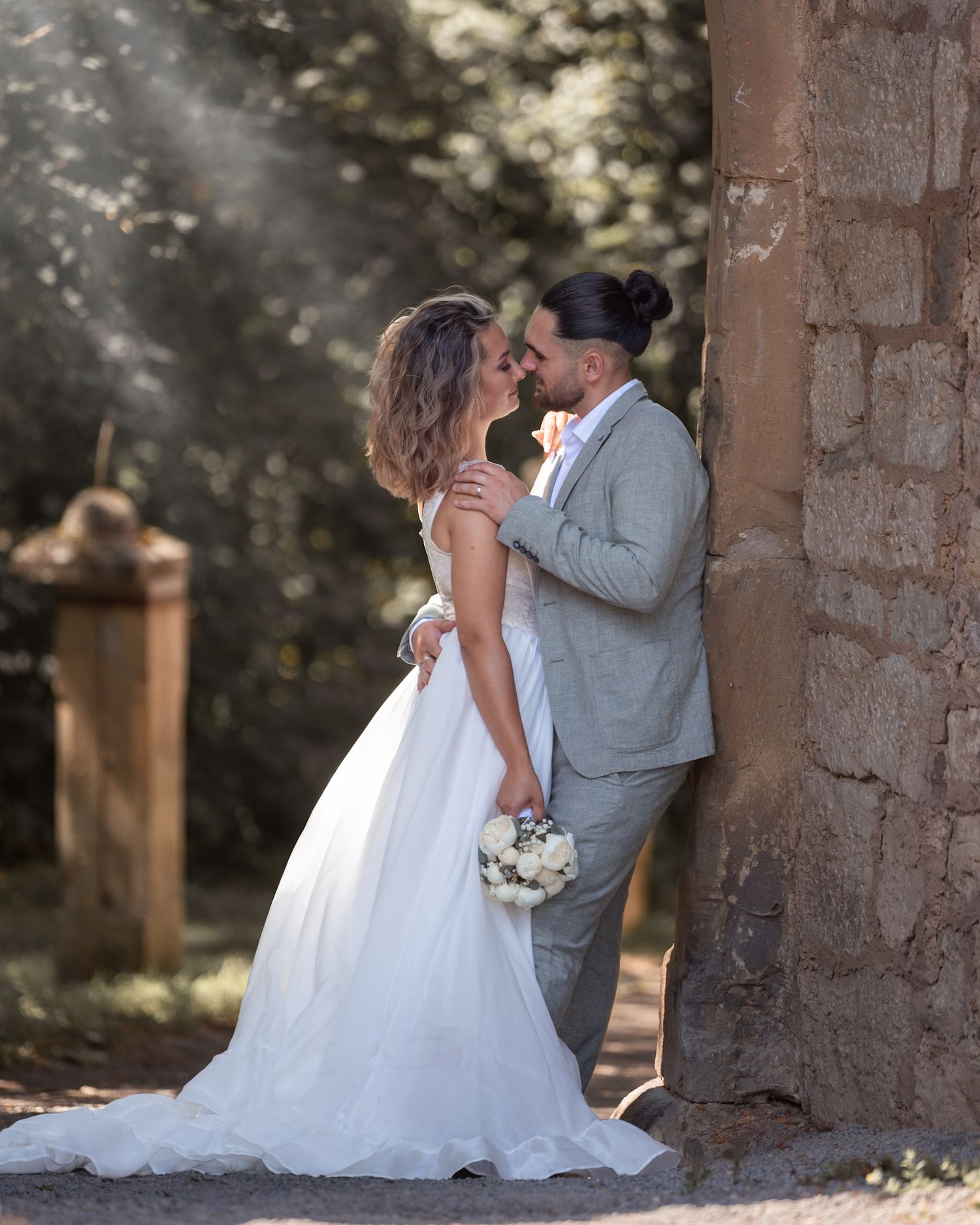Bride and groom share a romantic moment leaning against a stone wall, surrounded by lush greenery in soft natural light.