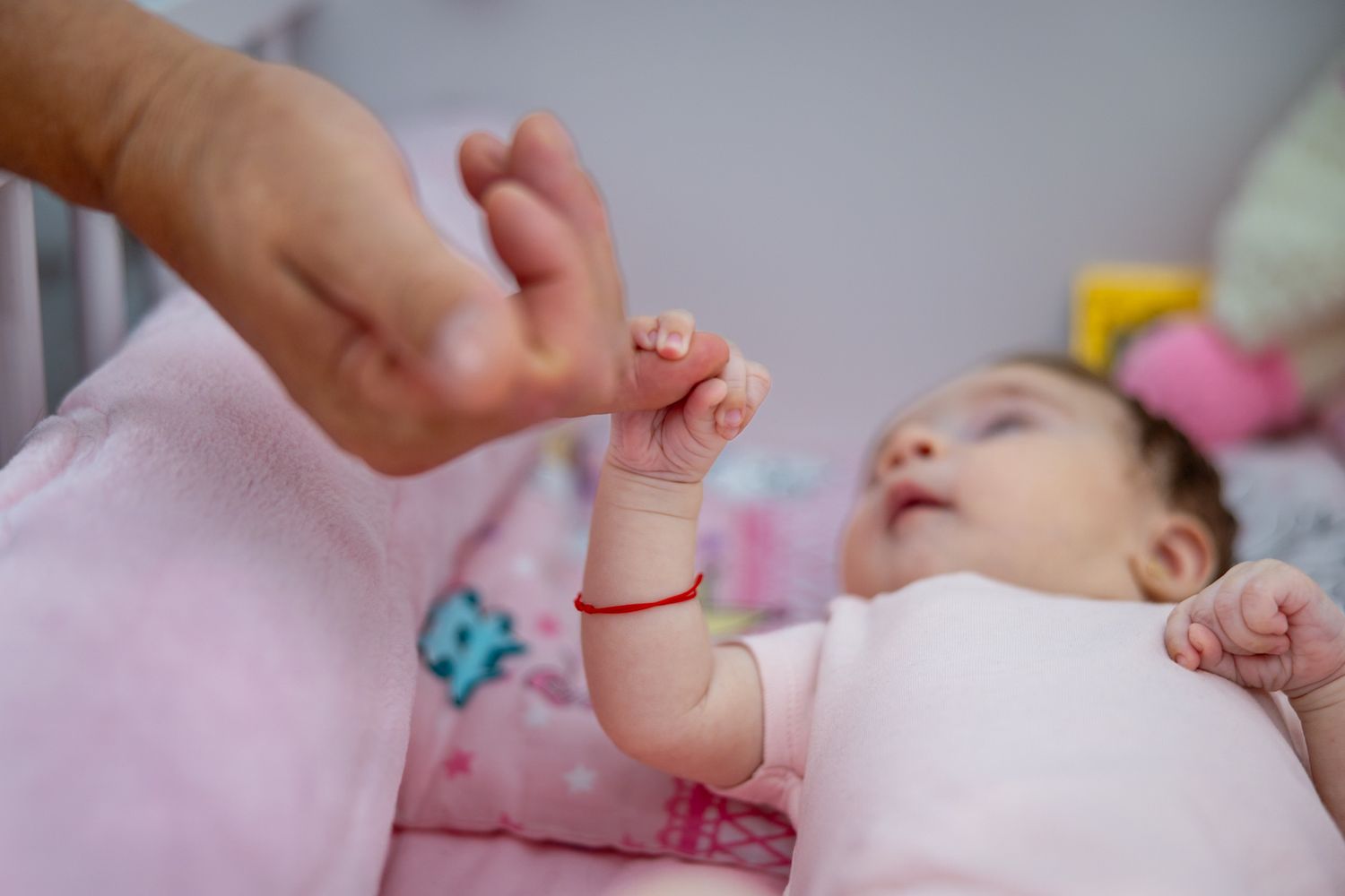 Newborn baby lying on pink bedding, grasping an adult finger with tiny hand.
