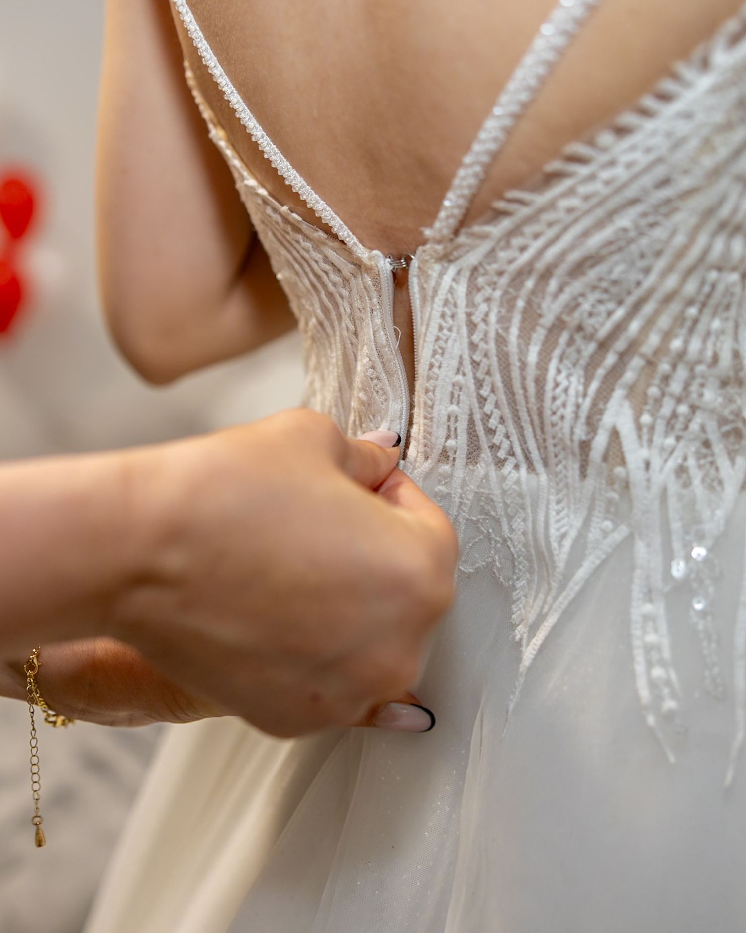 Hands fastening the back of a white lace wedding dress with intricate beaded details and delicate straps.
