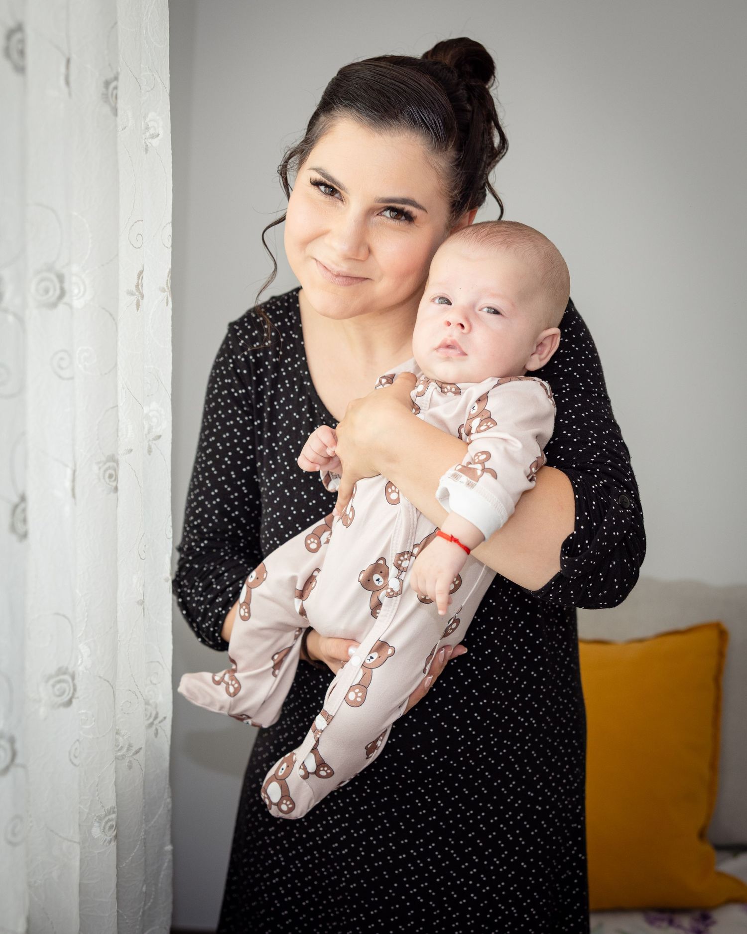 A smiling woman in a black dress holds her infant baby dressed in a cute bear-print onesie near a bright window.