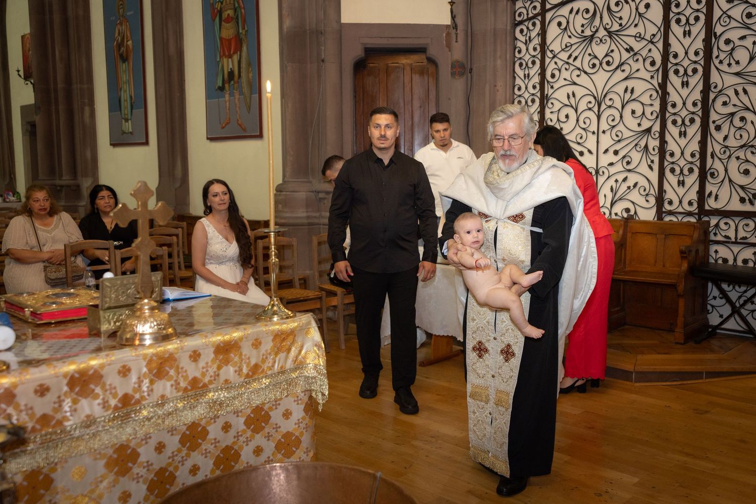 Priest holds infant during baptism ceremony in ornate church with guests seated in background.