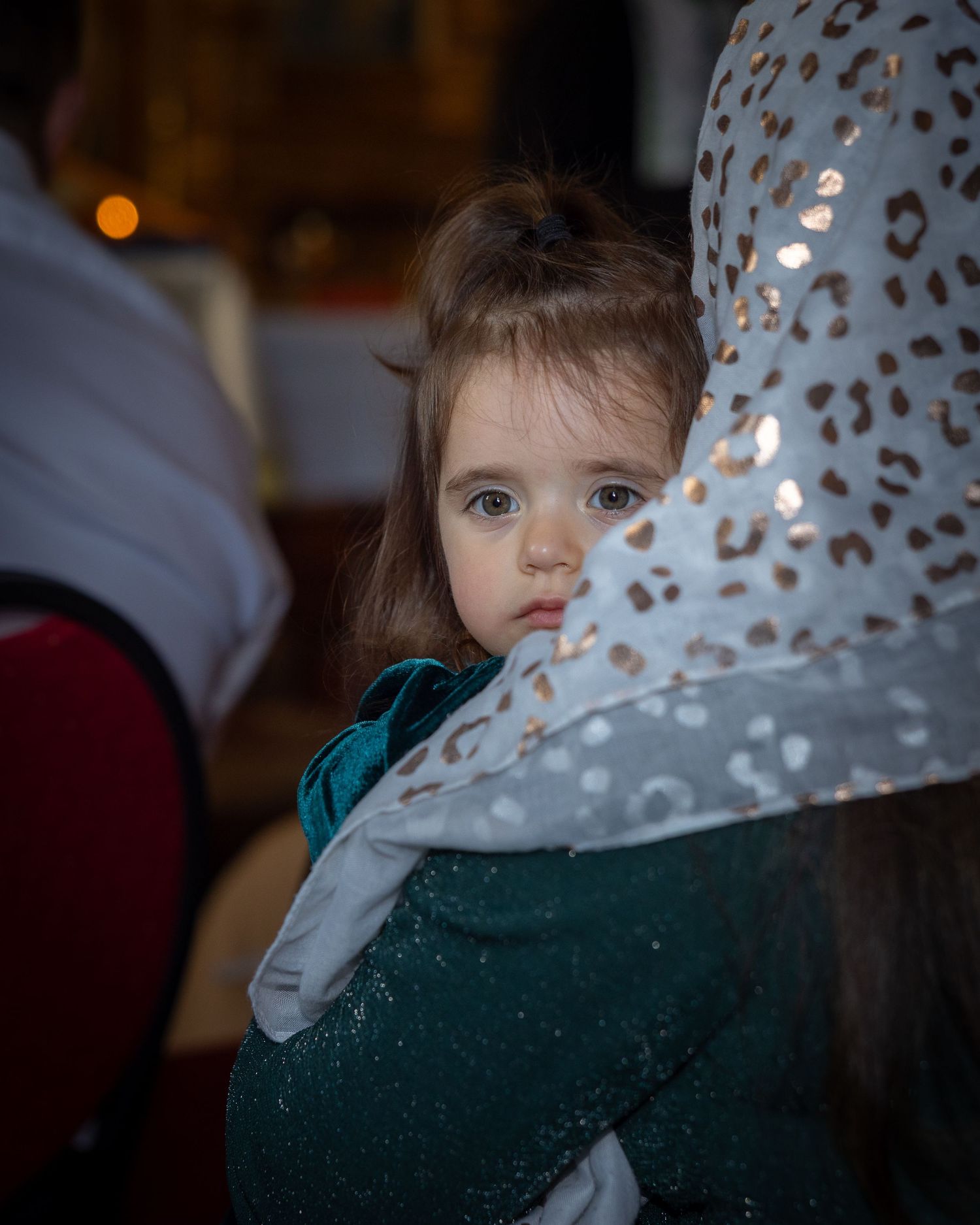 Ein kleines Mädchen im grünen Kleid blickt über die Schulter einer Frau mit gemustertem Kopftuch in die Kamera.