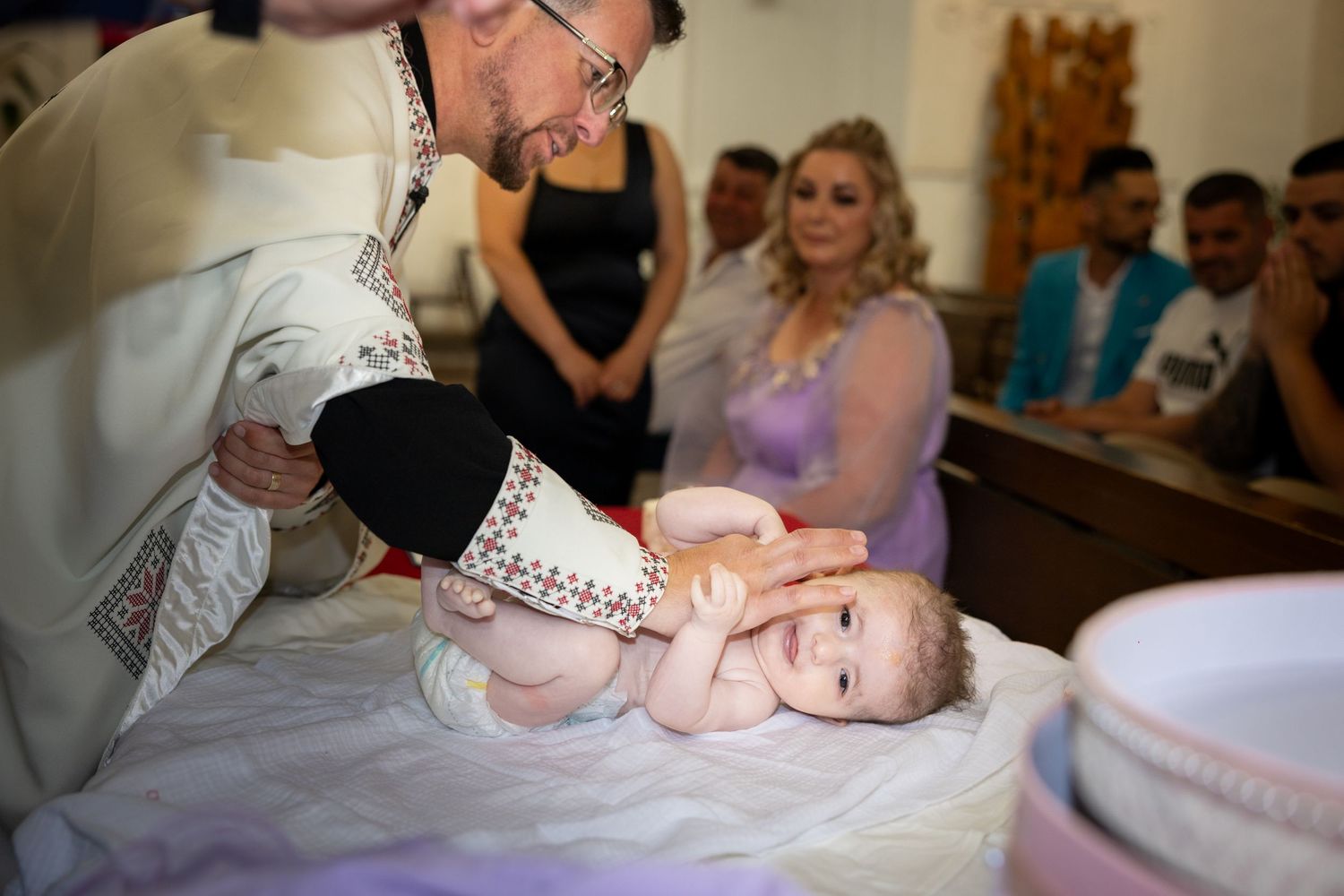 Priest performing a baptism ceremony, gently holding a baby on a white cloth near a baptismal font.