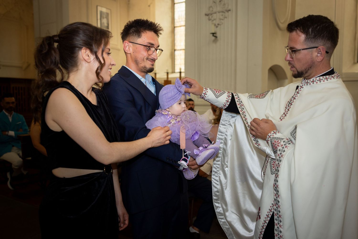 Priest performing a baptism ceremony on a baby dressed in purple, with parents smiling in a church.