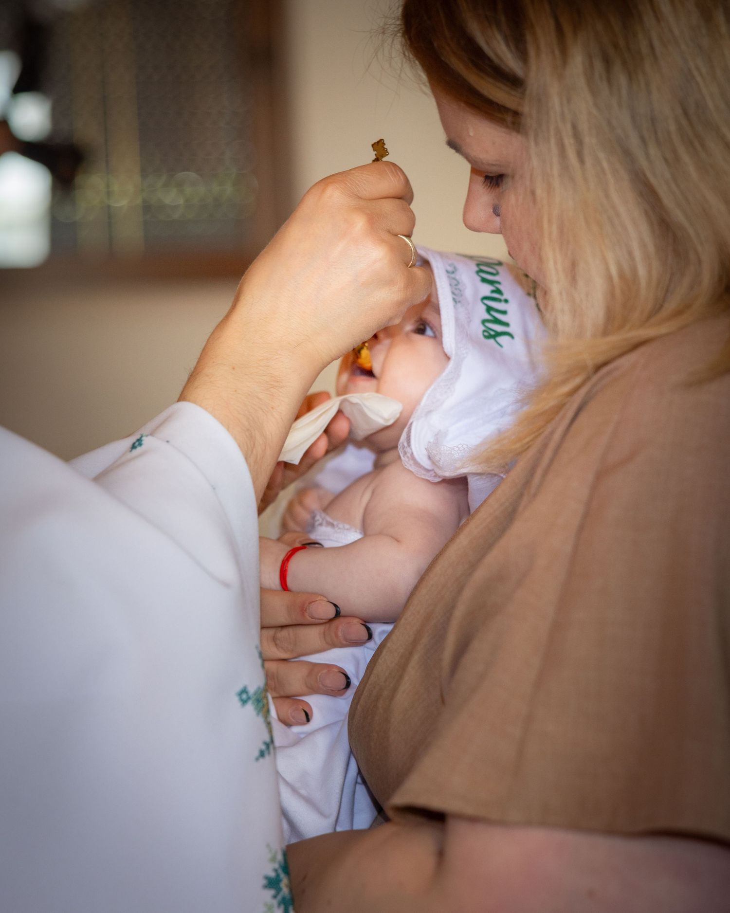 A mother holds her newborn baby during a religious baptism ceremony with a priest administering holy communion.