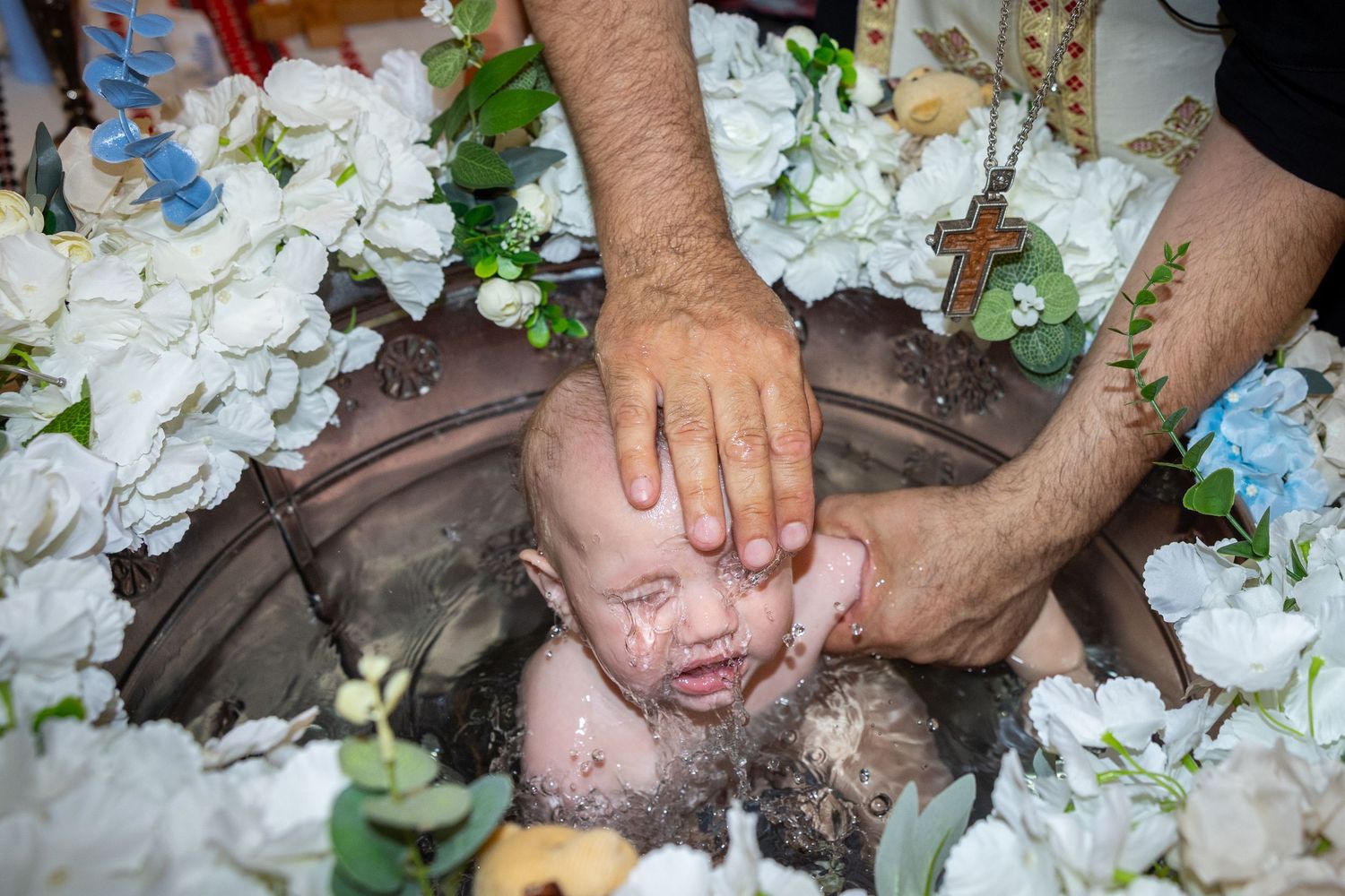 Der Priester hält Aris im blumengeschmückten Taufbecken, während Wasser über sein Gesicht läuft.