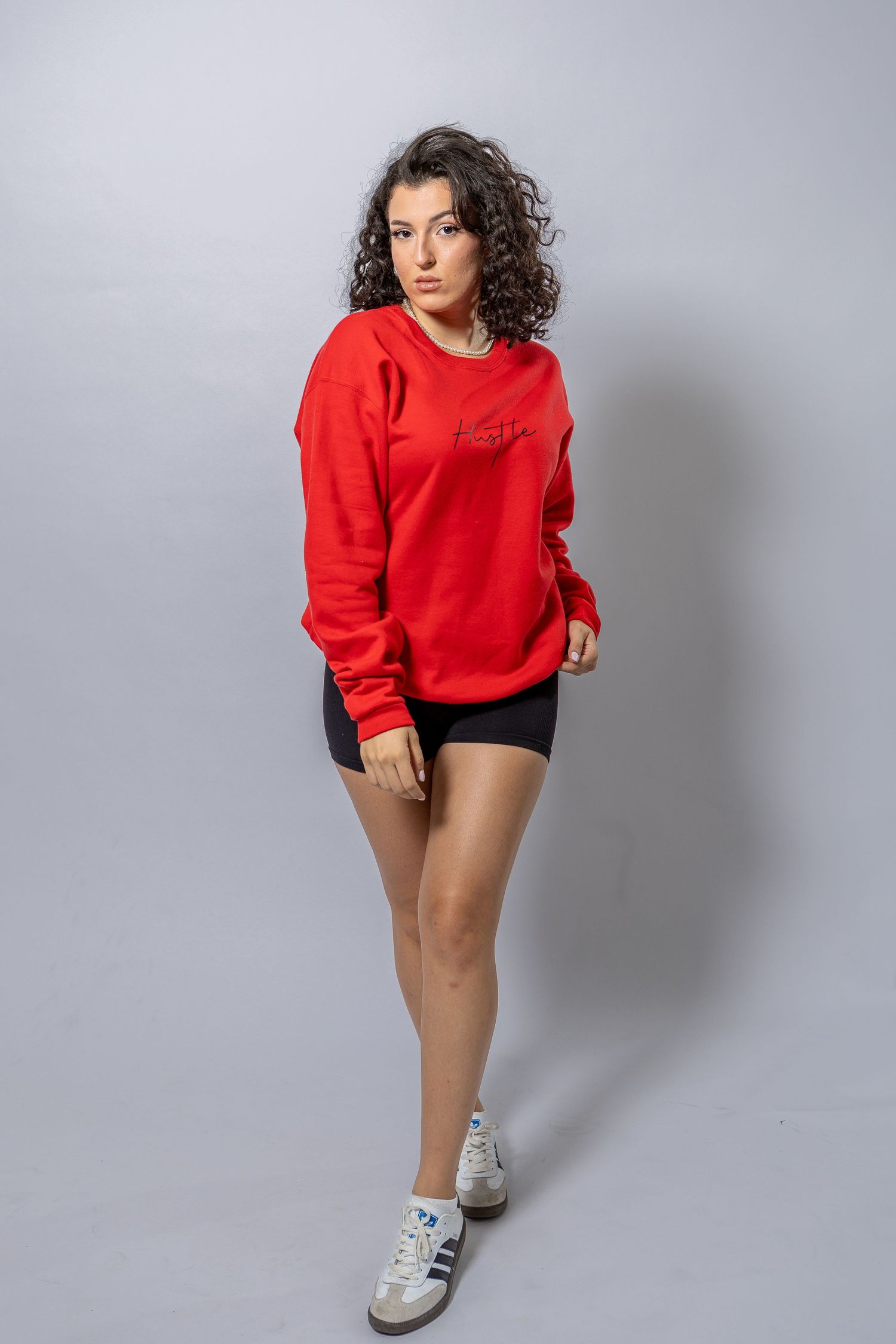Young woman with curly hair wearing a red oversized sweatshirt, black bike shorts, and white sneakers on a gray background.
