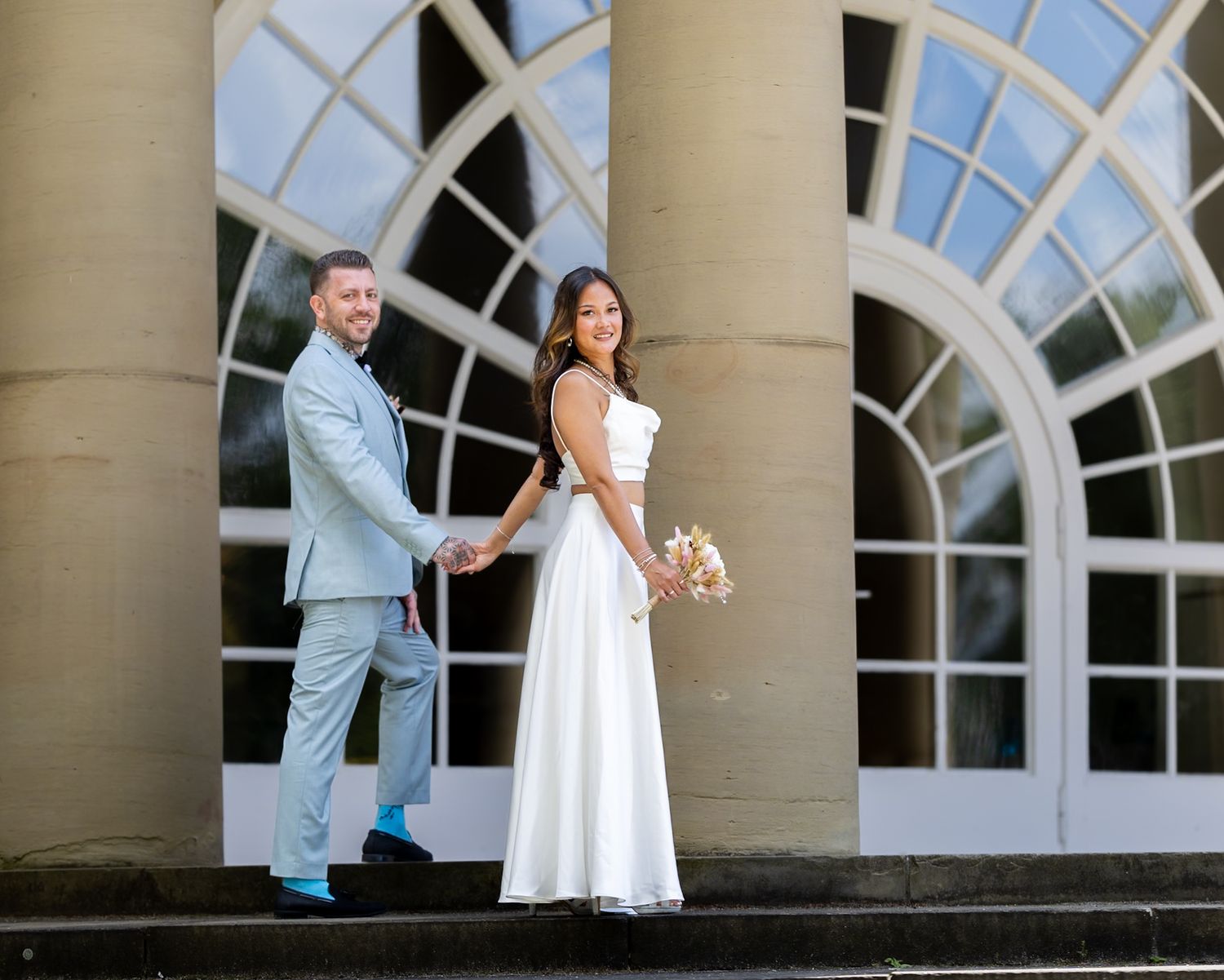 Bride and groom holding hands in front of elegant arched windows at their wedding venue.