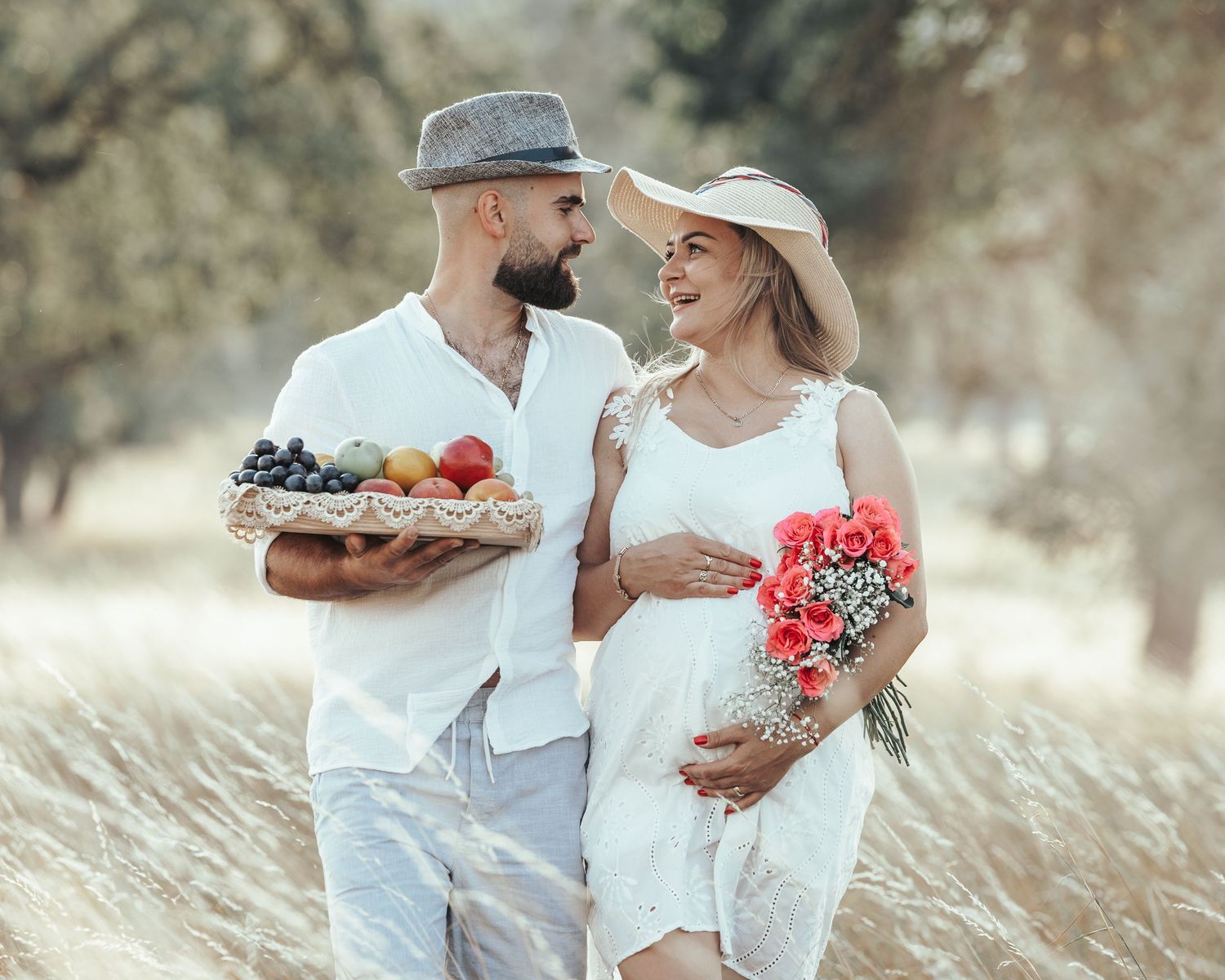 Pregnant couple in white outfits walking in field, man holding fruit basket, woman holding red flowers.