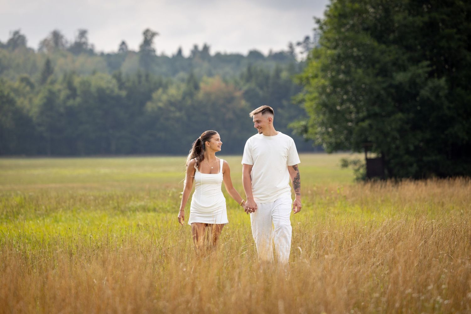 Weite Ansicht der Wiese: Lorena und Bogdan gehen Hand in Hand vor einer Baumreihe.