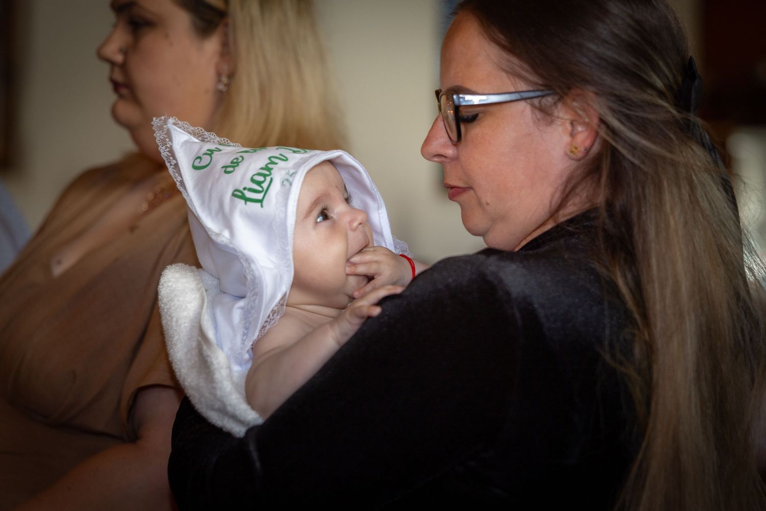 Woman holding a baby wrapped in a white hooded towel with green logo at an indoor gathering.