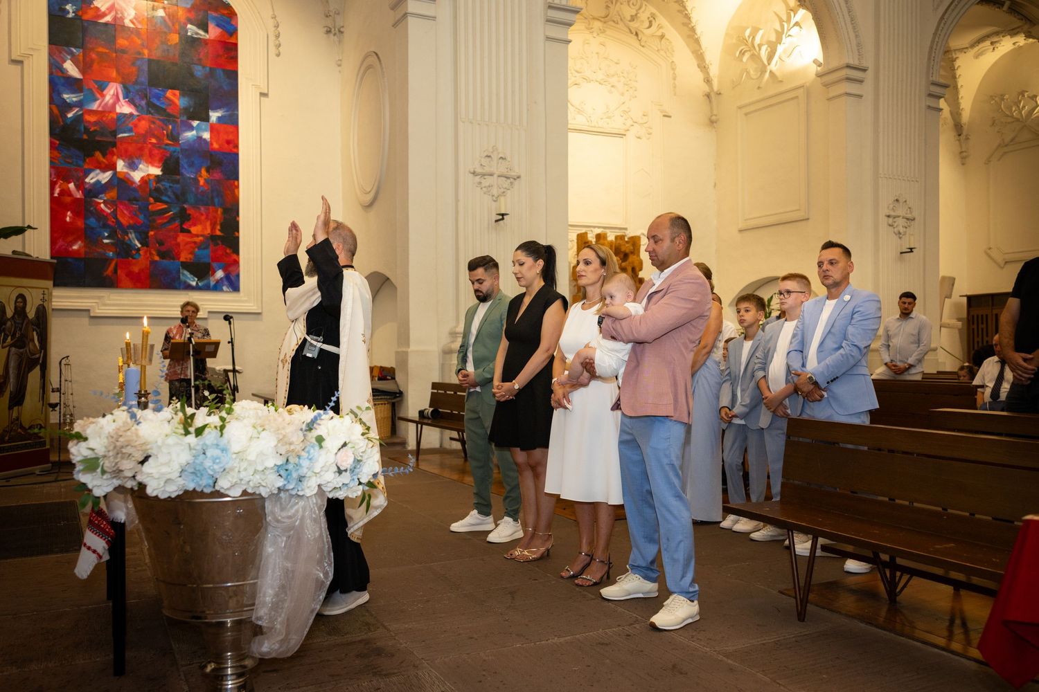 Der Priester erhebt die Hände am blumengeschmückten Taufbecken; Aris und die Taufgesellschaft stehen dahinter.