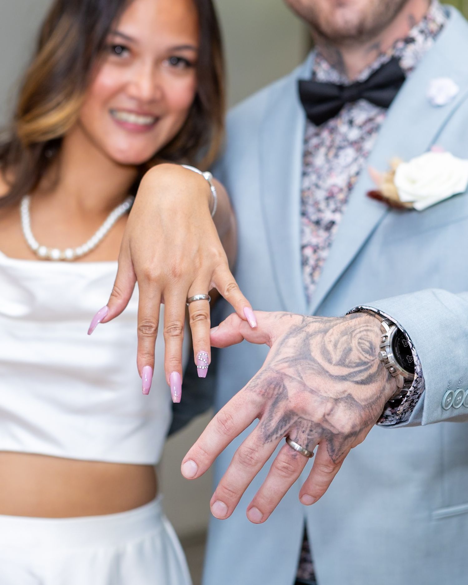 Newlyweds showing off wedding rings, bride with pink nails in white crop top, groom in light blue suit with rose hand tattoo.
