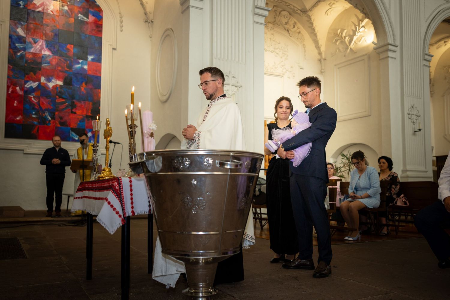 Alexia Ioana wird neben dem Priester und dem Taufbecken auf dem Arm gehalten, umgeben vom hellen Kirchenraum.