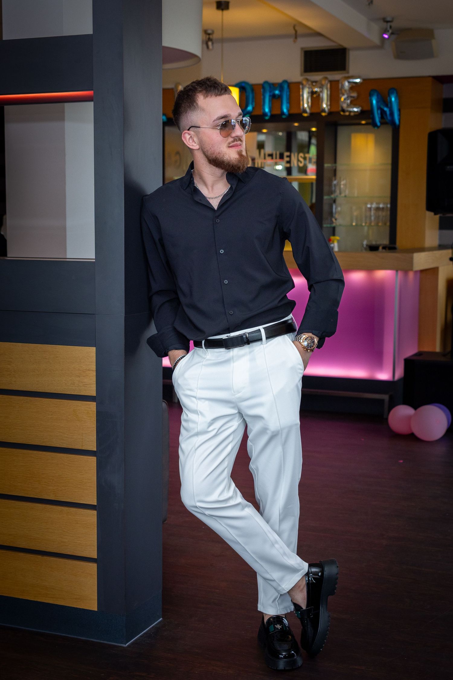 Mann mit Sonnenbrille, schwarzem Hemd und weißer Hose lehnt entspannt an einer Säule vor einer beleuchteten Bar.