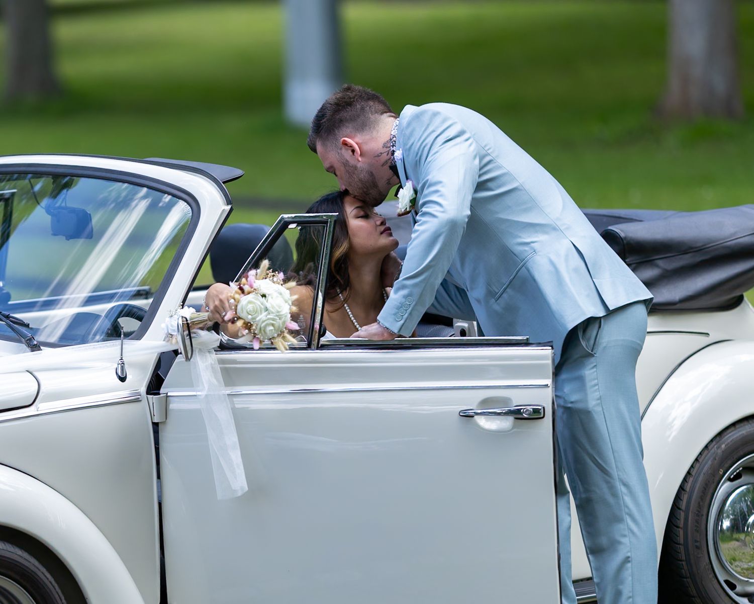 Groom in light blue suit kissing bride beside white vintage car decorated with flowers at outdoor wedding.