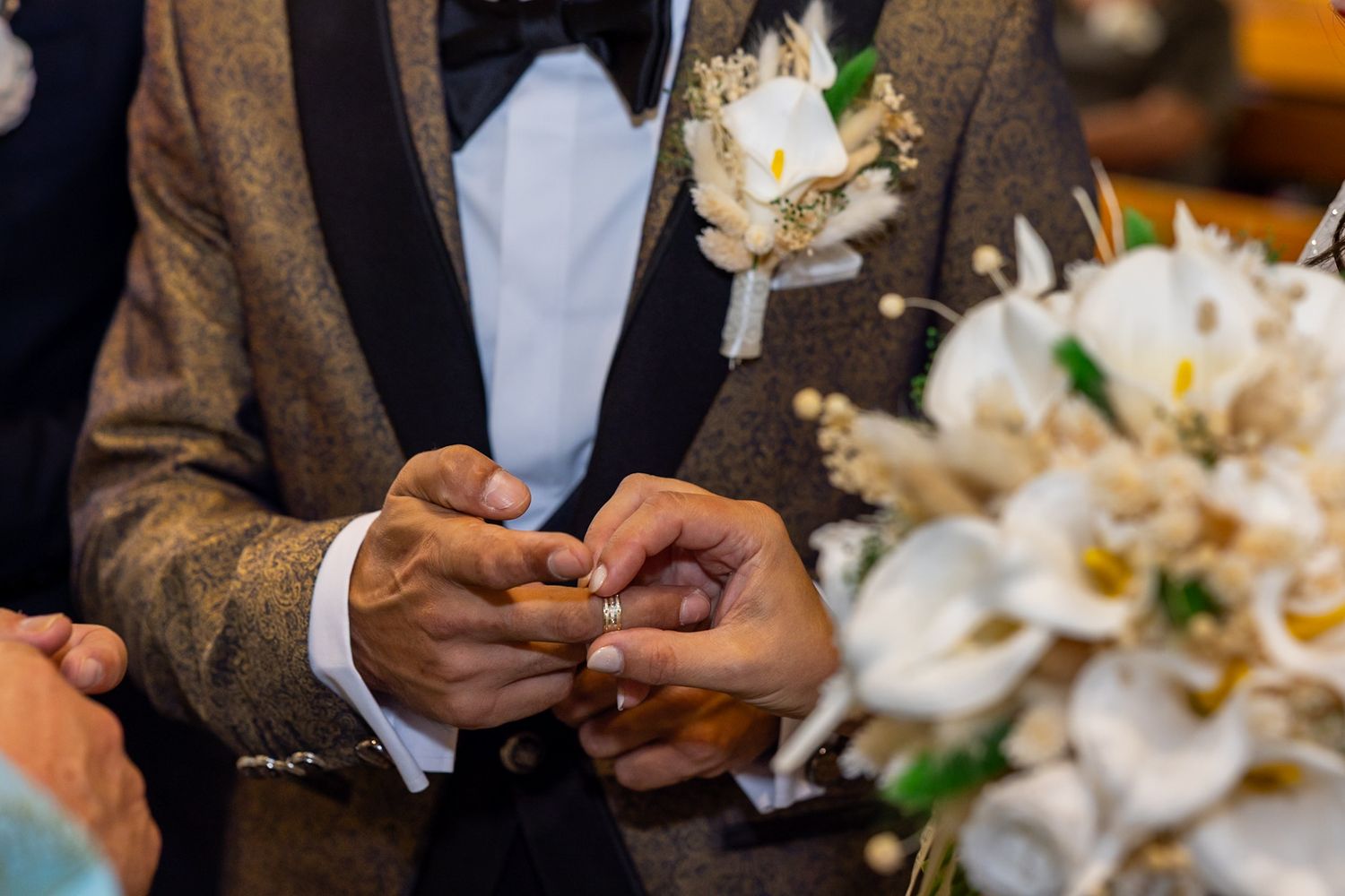 Groom placing wedding ring on bride's finger during ceremony with white floral bouquet visible.