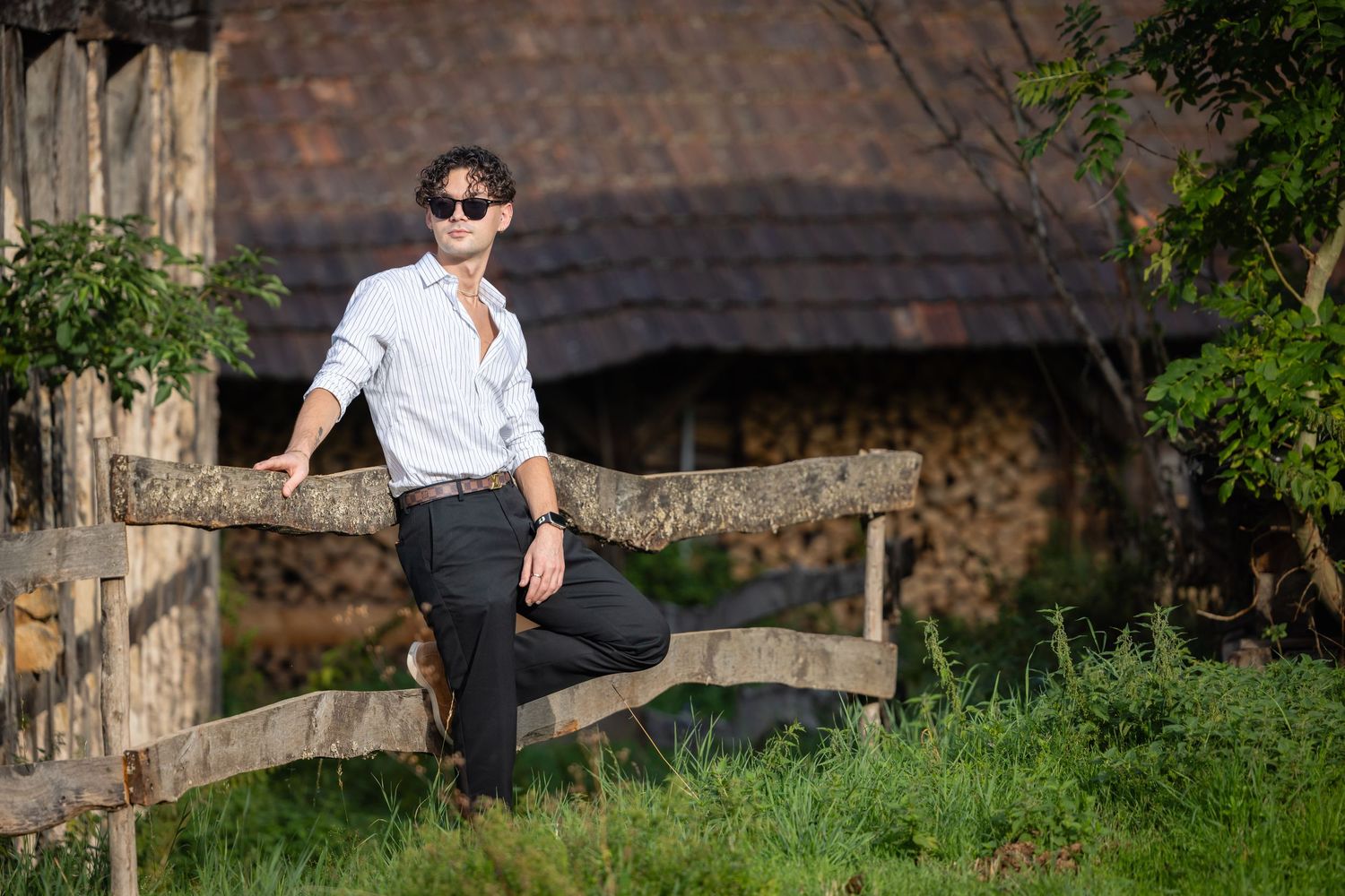 Young man in white shirt and sunglasses leaning on rustic wooden fence with countryside barn background.