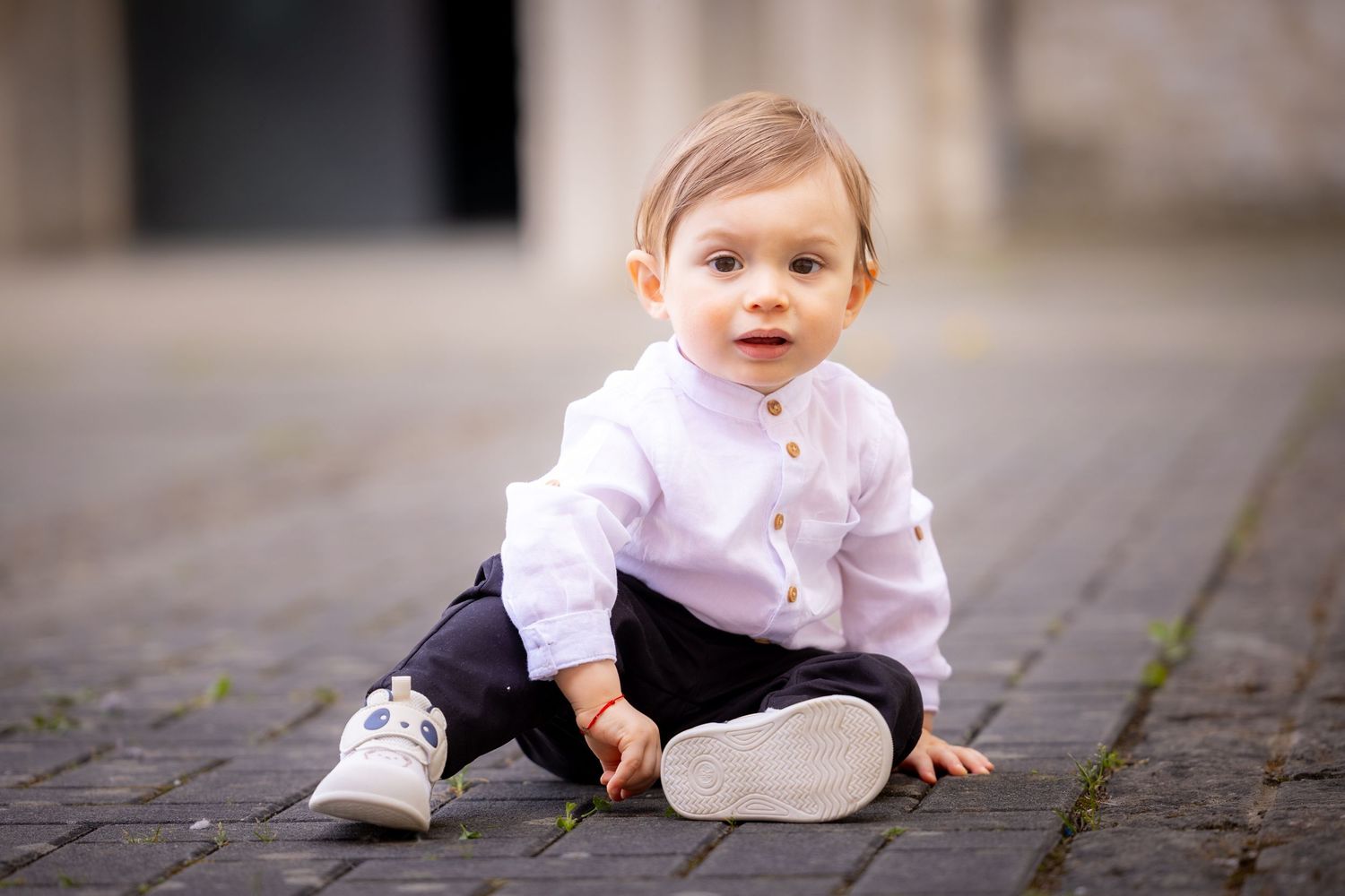 Adorable baby boy in white shirt and dark pants sitting on cobblestone pavement outdoors.