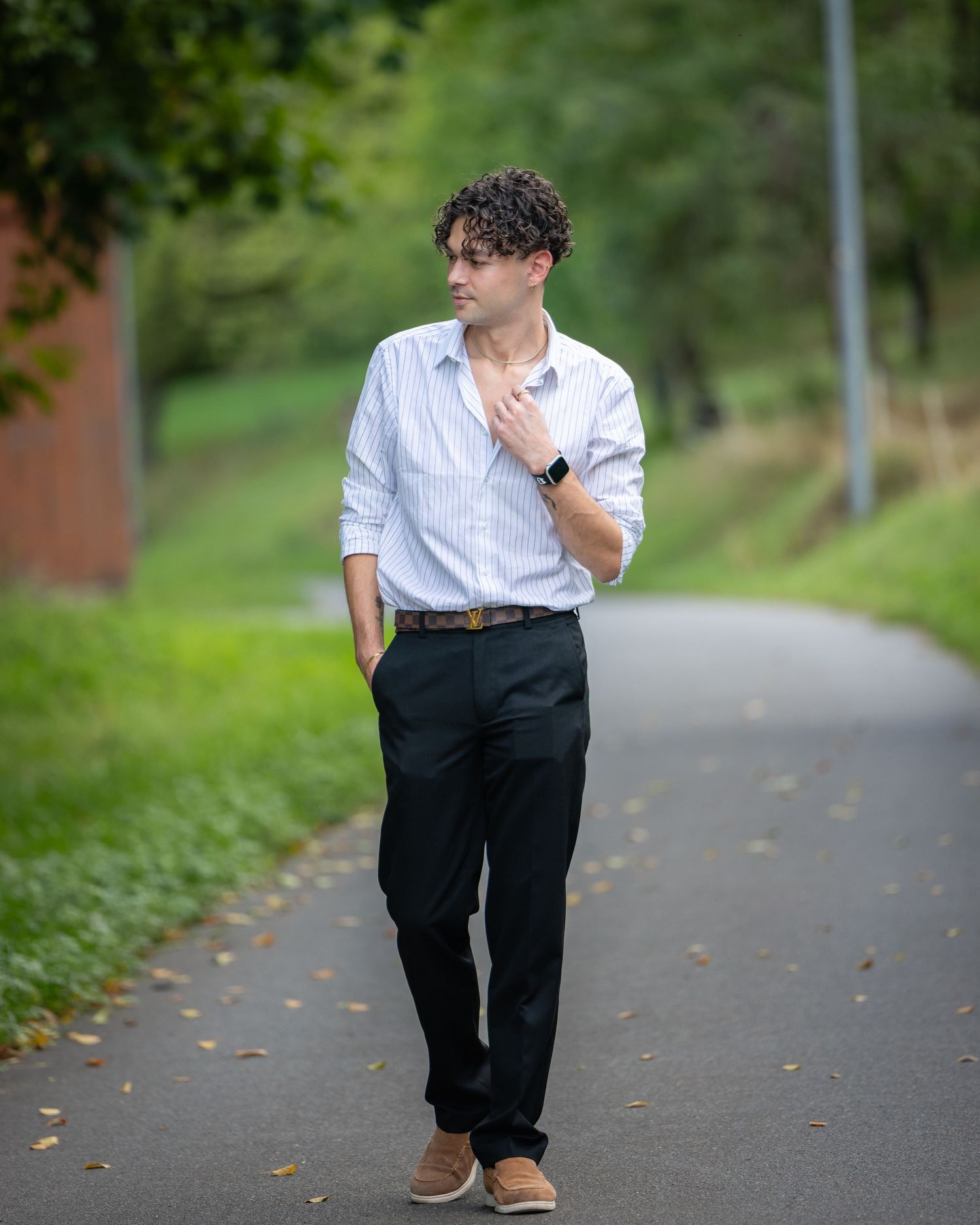 A young man in a striped shirt and black trousers walks confidently down a tree-lined path outdoors.