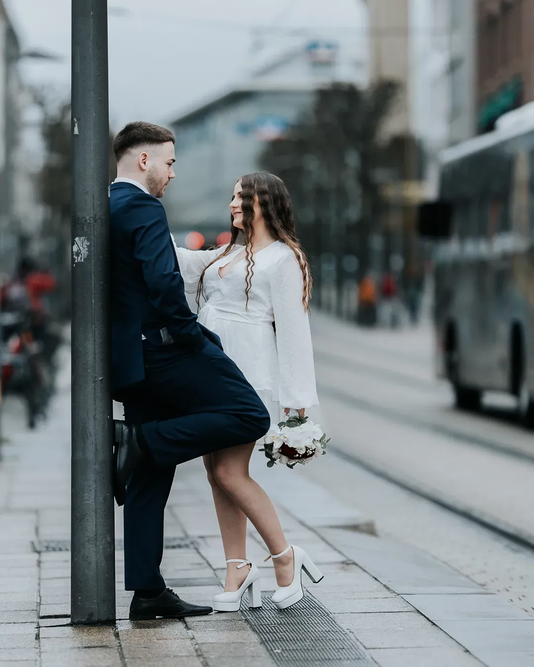 A newlywed couple shares a romantic moment at a city tram stop, bride in white mini dress holding a bouquet.