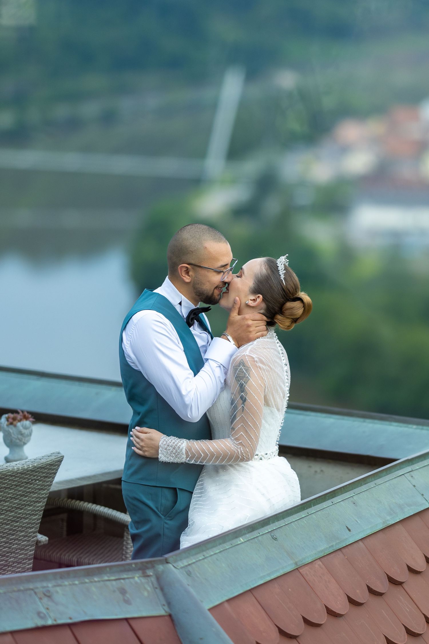 Bride and groom share romantic kiss on rooftop terrace with scenic river and green hills in background.