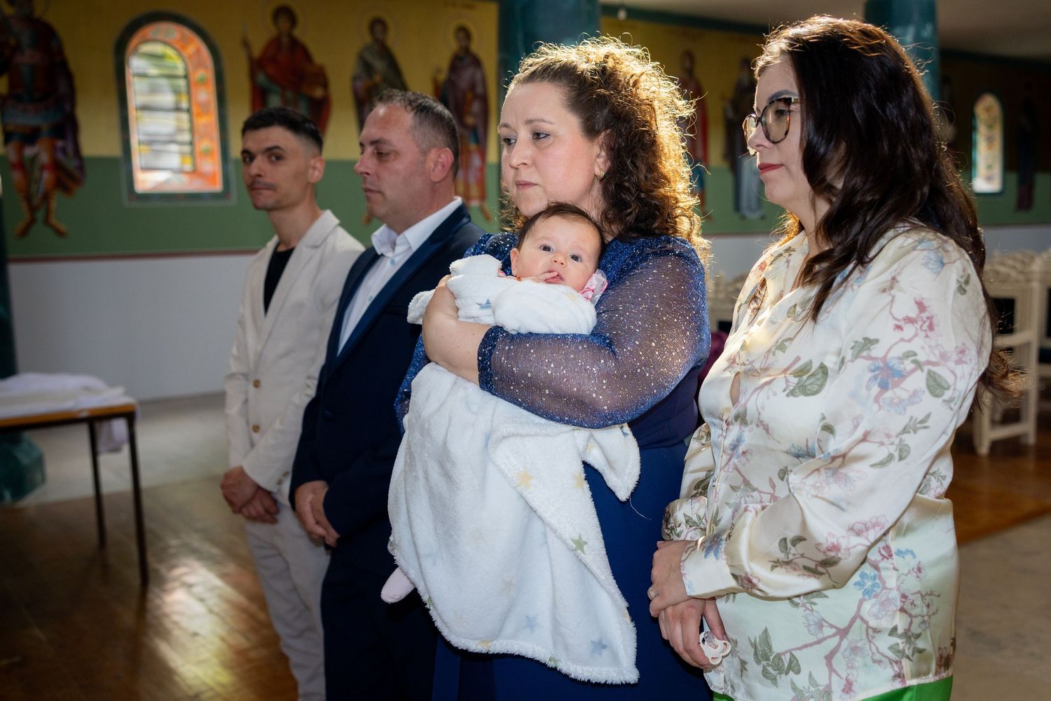 Family gathered in a church for a baptism ceremony, with a woman holding a baby dressed in white christening gown.