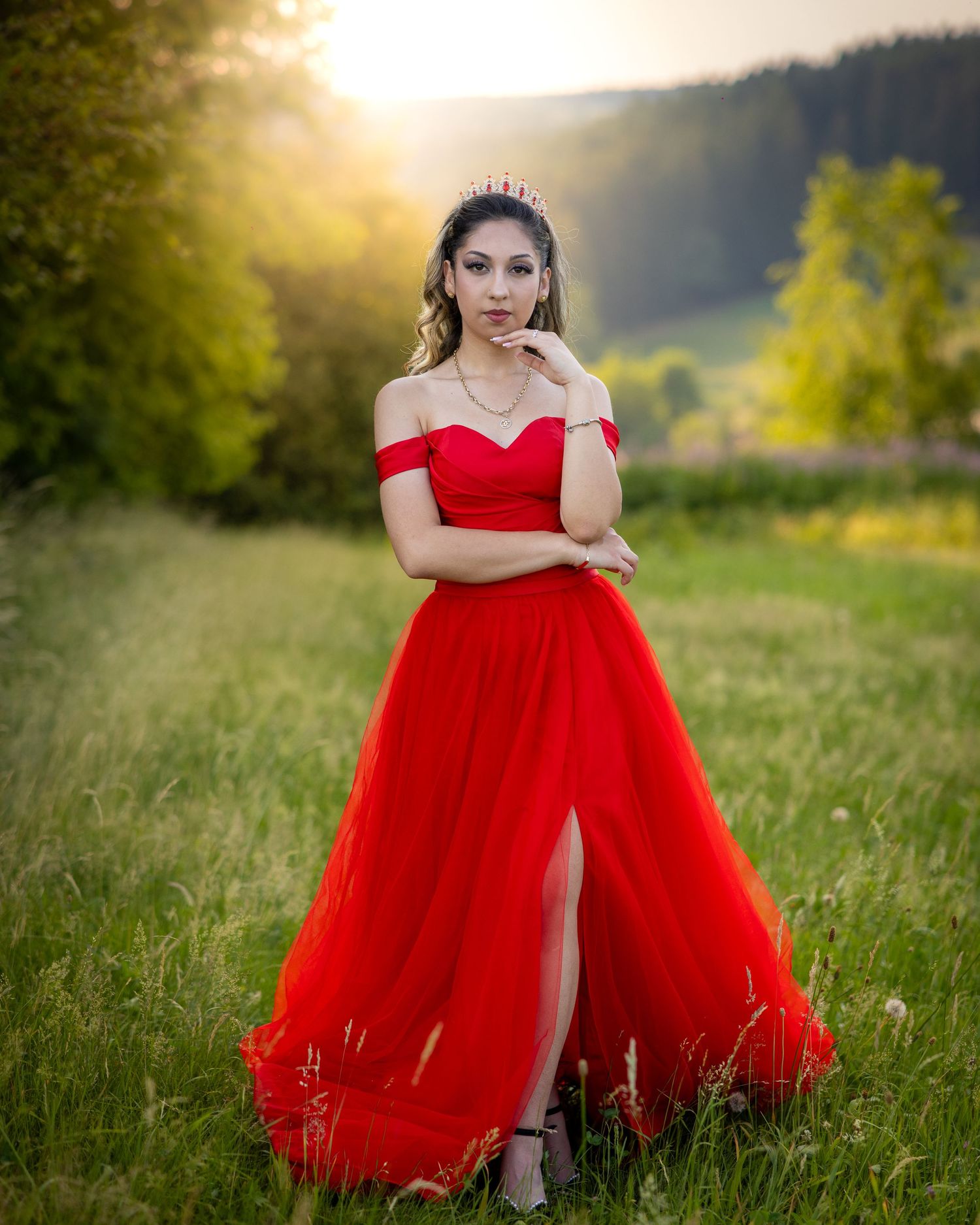Junge Frau im roten Abendkleid und mit Tiara auf einer Wiese im warmen Gegenlicht der Abendsonne.