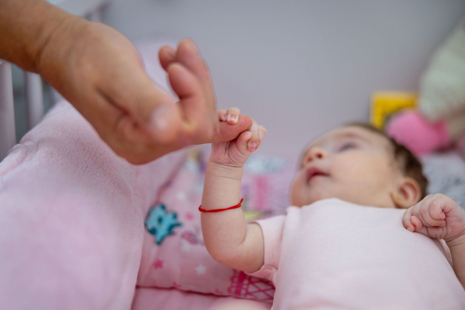 A newborn baby lying on a pink blanket in a crib, grasping an adult's finger with tiny hands.